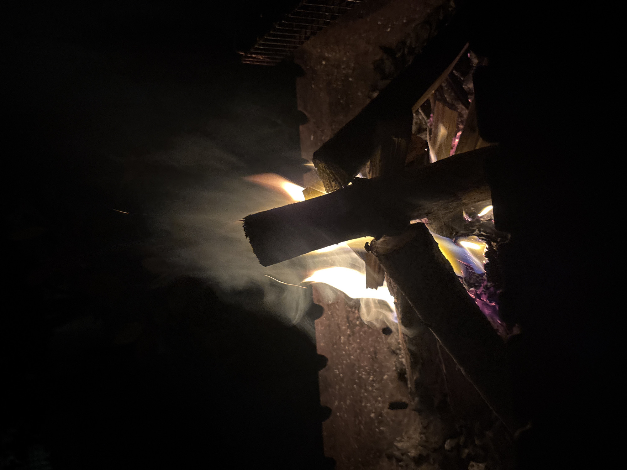 A campfire is burning in a metal brazier at night. The fire is made of logs and has flames and smoke. The brazier is filled with ashes and embers. The background is dark and out of focus. There are no people in the image. The location of this scene is unknown and cannot be determined from the image. It is not possible to determine if the scene is in Krasnodar, Russia.