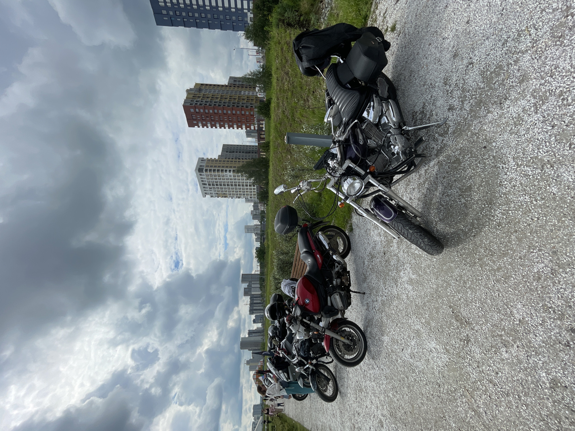 The image depicts a scene from the Solnechny (Солнечный) residential district in Yekaterinburg, Russia, on an overcast day.
In the foreground, a light grey gravel path occupies the bottom half of the frame. Several motorcycles are parked on this path, extending from the right foreground to the mid-left. The closest motorcycle, positioned on the right, is a dark purple/black Yamaha cruiser-style motorcycle with prominent chrome engine components and a black ribbed seat. A black leather jacket is draped over its rear seat and fender, which also features a black leather saddlebag on the right side. Its front wheel and fender are partially visible, angled left.
To the mid-left, a red BMW motorcycle with a black engine and seat is parked on its kickstand, facing right. A black helmet rests on its seat. Behind this BMW, two additional motorcycles are partially visible: a dark grey/black touring-style bike and another cruiser. A light wooden park bench is situated behind the red BMW and the other parked motorcycles.
On the far left of the gravel path, a small group of people is present. A woman with light hair, wearing a white long-sleeved shirt and light trousers, stands near one of the motorcycles, looking downwards. Behind her, other individuals, including a child and an adult, are seen further down the path, moving away from the camera.
The mid-ground features a grassy embankment running parallel to the gravel path on its right side. Sparse green vegetation, including small bushes and nascent trees, dots this embankment. A light grey metallic pole stands vertically on the grassy area behind the closest purple motorcycle.
The background is dominated by a modern residential complex. From left to right, visible structures include a white multi-story building, a distinctive tall red and yellow multi-story building, and a prominent dark blue multi-story building with a grid pattern of windows. Other less distinct high-rise buildings are visible further in the distance. The overall impression is of a developing urban area with new residential towers.
The sky is heavily overcast with a multitude of grey clouds, ranging from lighter, textured formations to darker, denser masses. Patches of brighter, whiter sky are visible through the clouds, particularly in the upper left quadrant, and hints of blue sky are present in isolated spots.