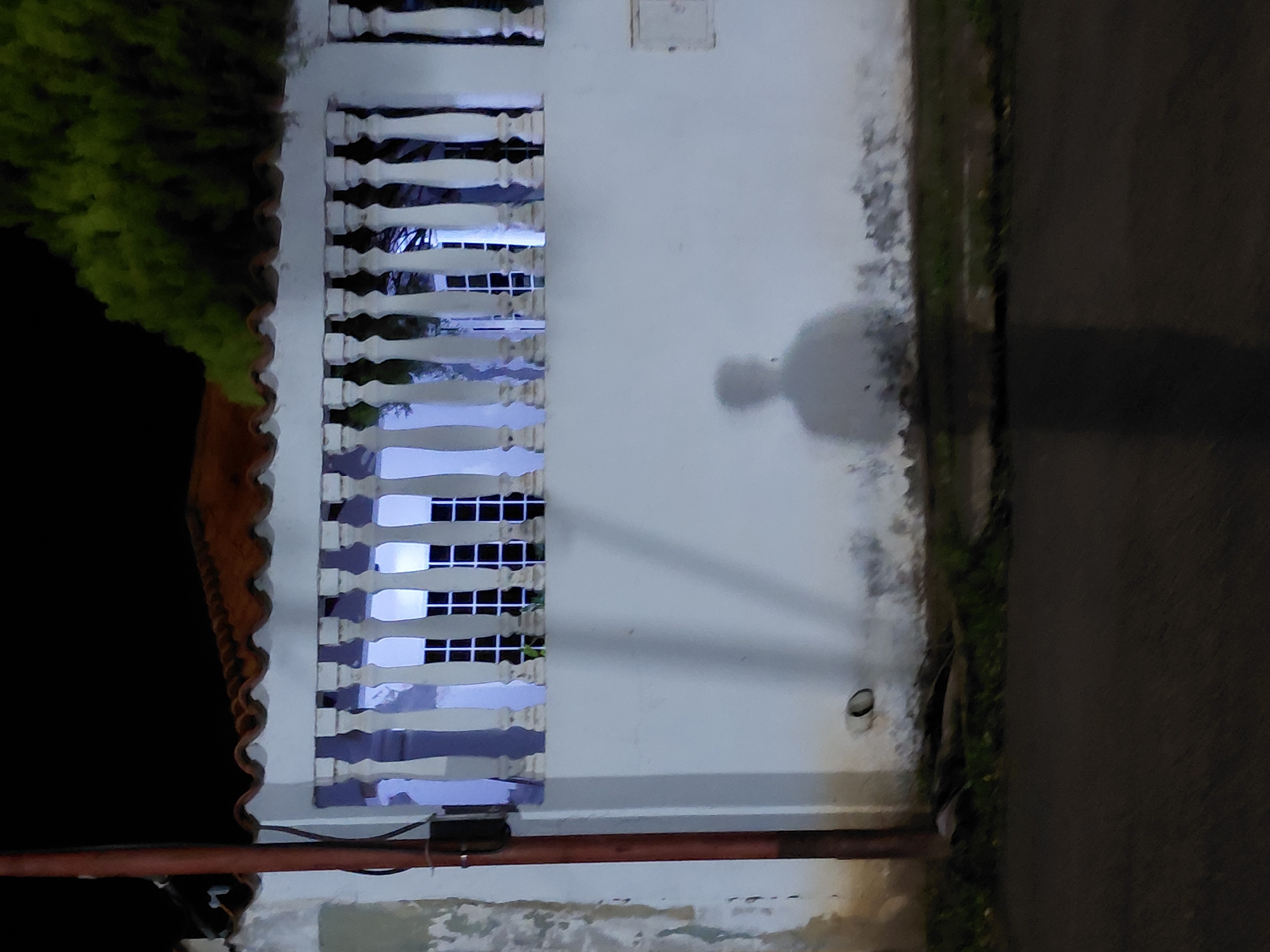 The image captures a nighttime scene in Santa Ana, Venezuela, focusing on a white stucco wall with a decorative balustrade running across its upper portion. Behind the balustrade, a window with a grid pattern is visible, illuminated by a soft blue light, suggesting interior lighting. To the left, a reddish-brown pole, possibly a utility pole, stands vertically, attached to which is a dark electrical box. The wall itself shows signs of age and wear, with some peeling paint and staining near the base.
A prominent feature of the scene is a faint, silhouetted shadow of a person cast on the wall, positioned in the lower half of the frame. The shadow appears to be of a person standing, with a distinct head and shoulder shape, though lacking detailed features. Below the wall, a narrow strip of green grass and dark ground is visible, leading to a paved surface in the foreground, which could be a road or sidewalk. The overall atmosphere is somewhat muted and still, characteristic of a quiet night.
