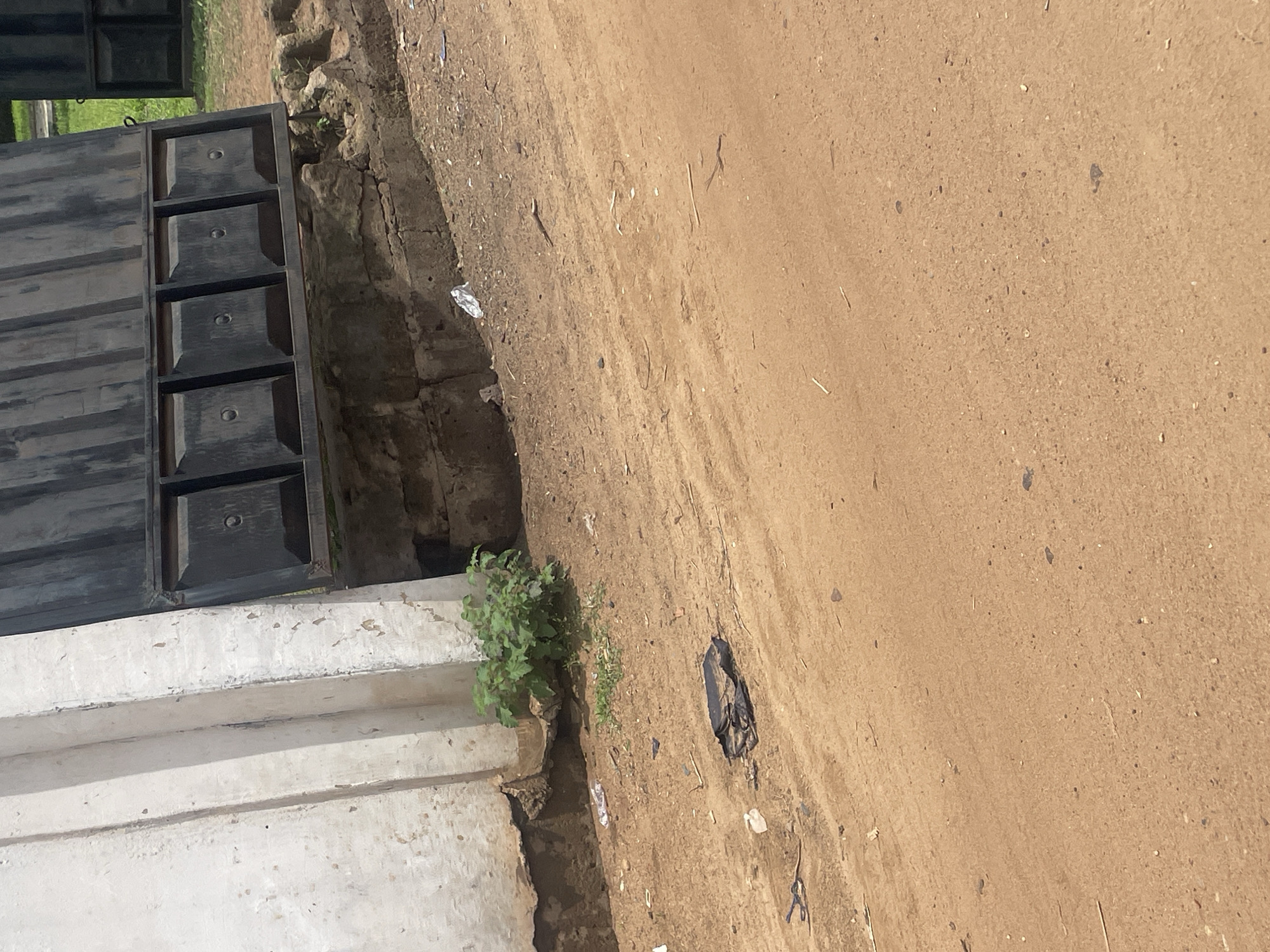 In the city of Lokoja, Nigeria, a dirt road runs alongside a partially constructed or aged wall. A large, dark metal gate, possibly part of a larger structure, is visible at the top of the frame, supported by a brick foundation. The gate has a series of rectangular panels, each with a circular indentation. Below the gate, the ground is a mix of dirt and rough stones. To the left of the gate, the wall is painted white and shows signs of weathering and chipping. A small cluster of green plants grows at the base of the wall. A black plastic bag and some other debris are scattered on the dirt road in the foreground. The scene suggests an outdoor, possibly semi-abandoned or utilitarian area, with elements of both construction and neglect.