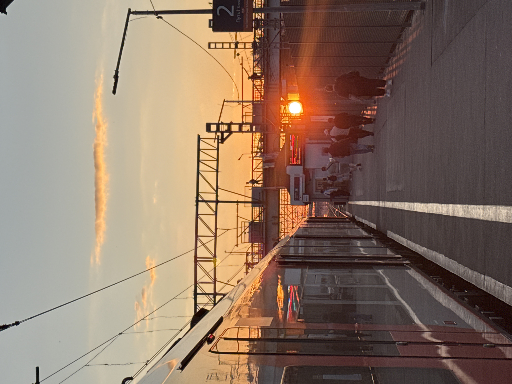 The image captures a sunset at a train station in Khimki, Russia. The sun, a bright orb, is low in the sky, casting a warm, orange glow over the scene and creating lens flare. A train is parked alongside the platform, its reflective surface mirroring the fiery sky and the overhead structures of the train tracks. Several people are visible on the platform, some walking, others standing, seemingly waiting for or disembarking from the train. The overall atmosphere appears calm, with the soft light of dusk lending a serene quality to the urban setting. Overhead train wires and station infrastructure are prominent features, indicating an active railway environment. The sky is a gradient of pale blue and warm orange, with a wispy cloud formation stretched across it. A sign with the number "2" and text in Russian, "Путь | Track", is visible, suggesting this is platform 2. The weather appears clear, with no indications of rain or strong winds. The light suggests it is late afternoon or early evening.