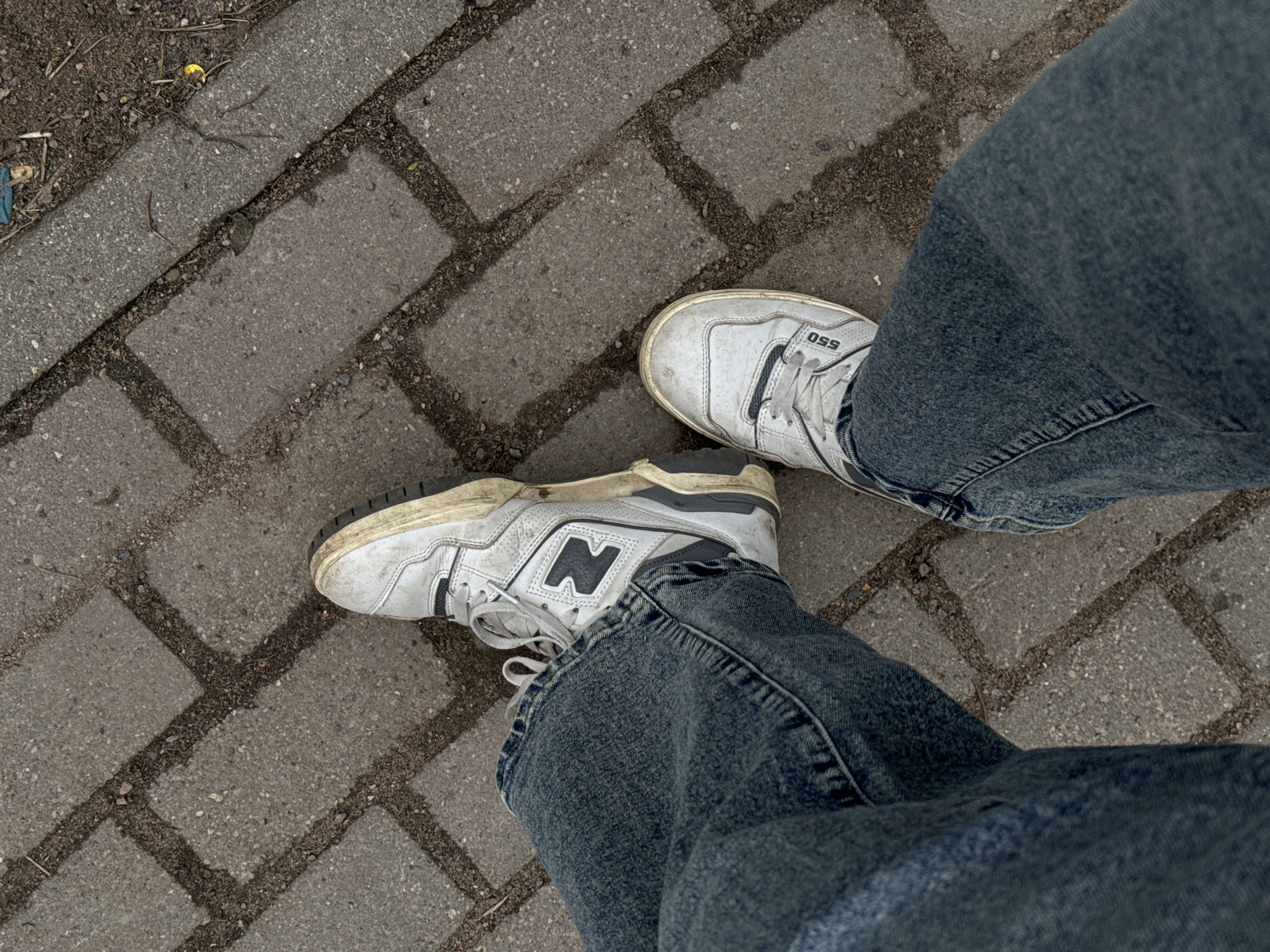 An eye-level, overhead shot depicts a person's lower body standing on a paved surface composed of rectangular blocks. The person wears loose-fitting, medium-wash blue jeans and white New Balance 550 sneakers, which appear worn. The paved ground is gray, with darker material filling the seams between the blocks. The location is Odintsovo, Russia.