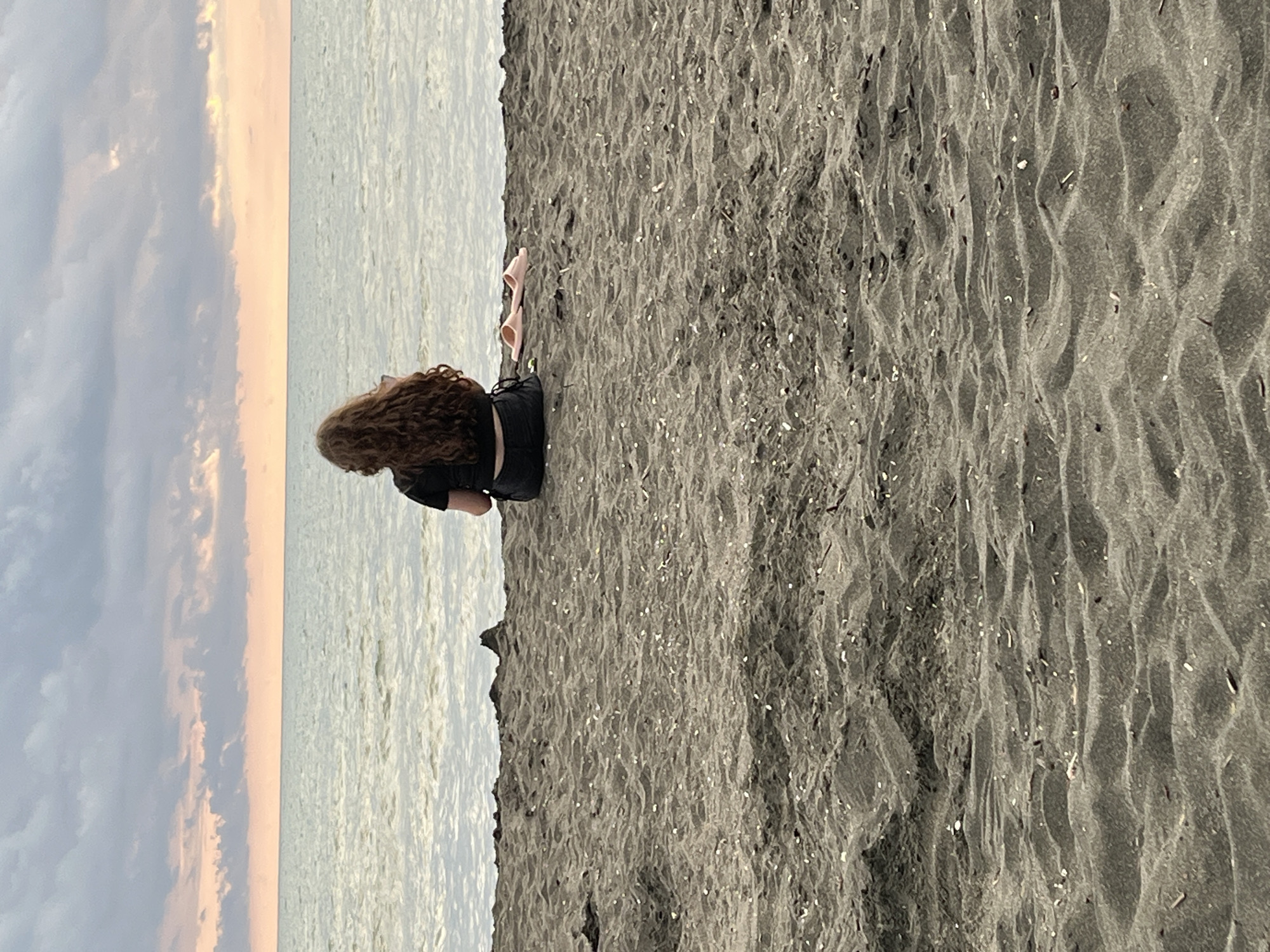 A person with long, curly brown hair is sitting on a dark sand beach, facing the sea. They are wearing a black t-shirt and dark shorts. In front of them, to their right, are two pink flip-flops. The sea is calm with small waves, and the sky above is filled with dramatic clouds with streaks of pink and orange from the setting sun. The sand is textured with small shells and debris. The scene is set in Ureki, Georgia.