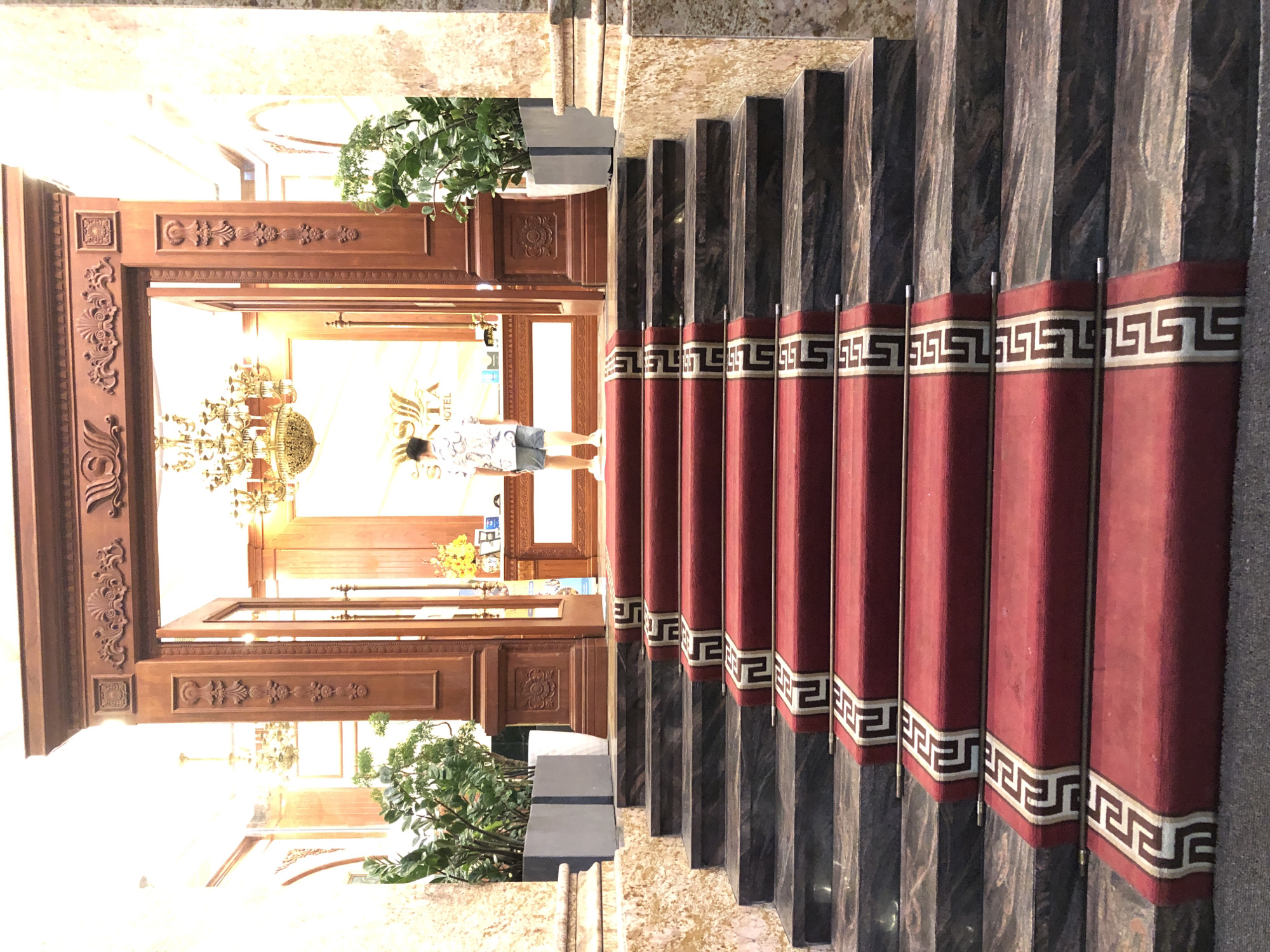The image depicts the entrance of a hotel in Da Nang, Vietnam. A person, viewed from behind, is walking up a grand staircase adorned with a red carpet and decorative borders. The staircase leads to a large wooden entryway with ornate carvings. Through the open doors, a reception area is visible, featuring a chandelier, plants, and a reception desk with a hotel logo that appears to read "SENTA HOTEL". The architecture suggests a luxurious establishment.