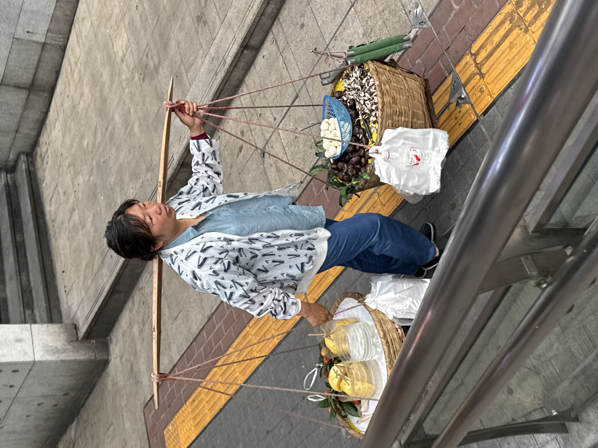 A woman walks along a Yuzhong District, China sidewalk carrying two baskets suspended from a pole across her shoulders. One basket contains what appear to be chestnuts, white balls, and plastic water bottles. The other basket contains fruit inside plastic containers. The woman wears a white patterned jacket, a blue shirt, and blue jeans. She is near a set of stairs and a metal railing. The sidewalk features both yellow and gray paving stones.