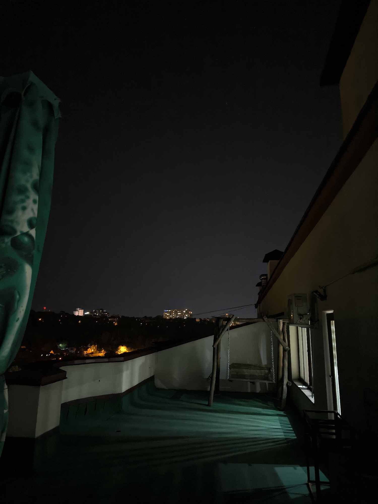 The image depicts a night scene from a rooftop overlooking a cityscape. The foreground features a dark green rooftop with distinctive shadow patterns cast by unseen light sources. To the left, a large, green, possibly fabric-covered object, likely an umbrella or canopy, is partially visible and creates a strong vertical element. In the mid-ground, there's a white parapet wall bordering the rooftop. A wooden swing set with a wooden seat hangs from chains, positioned near the edge of the rooftop. To the right, a building is visible with a section of its exterior wall, a window, and an air conditioning unit. Beyond the rooftop, the cityscape of Chișinău, Moldova, is visible with illuminated buildings and lights scattered across a dark, hilly landscape under a starless night sky. The overall impression is one of a quiet, urban night.