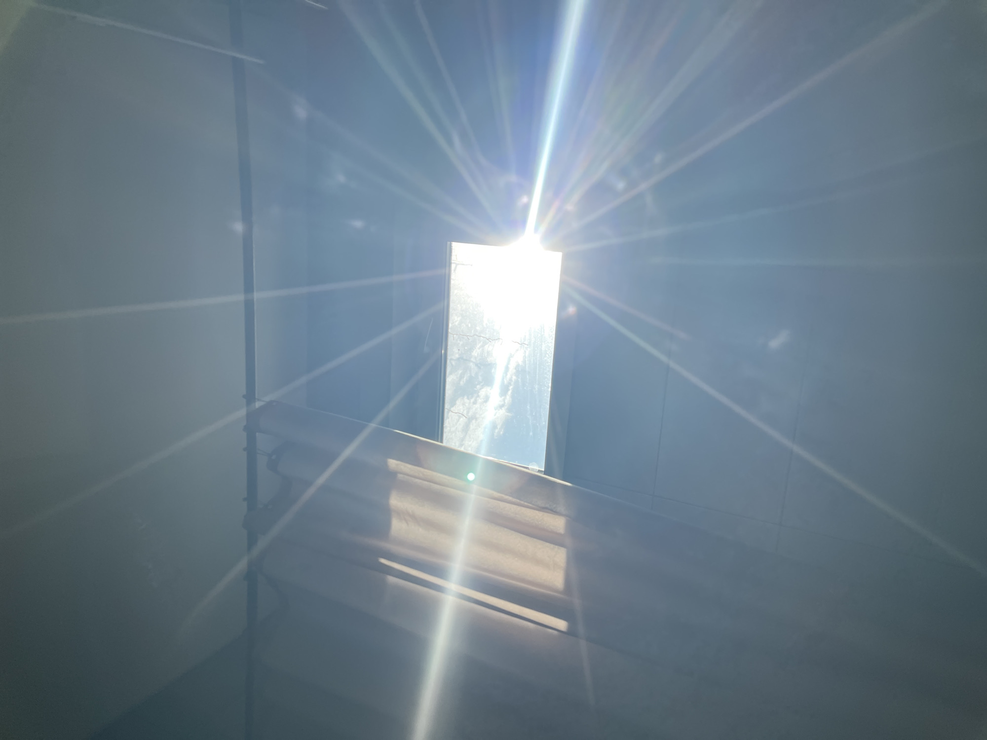 A photograph depicts a bright, rectangular skylight viewed from below. Intense sunlight streams through the opening, creating a starburst effect. The walls surrounding the skylight appear light blue. A window shade with visible slats is positioned below the skylight, partially obstructing the view. Based on the provided location, the image was likely taken in a building in Мосметаллоконструкция Онт, Russia.