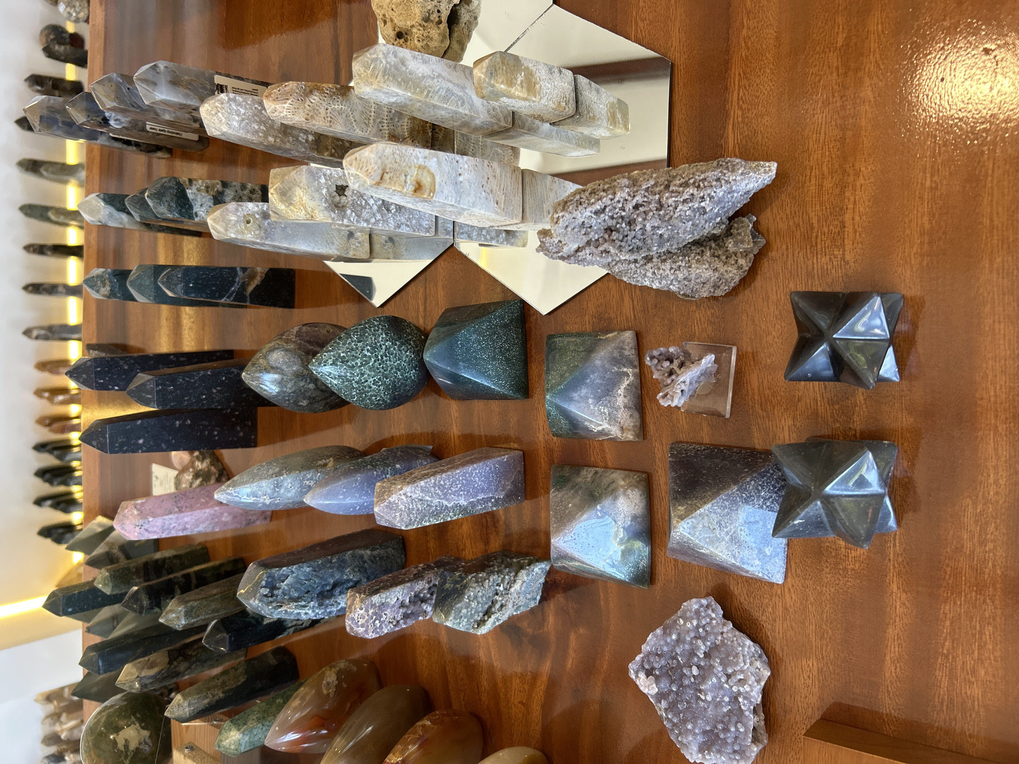 The image depicts a collection of polished stones and crystals displayed on a polished wooden surface. The items are arranged in rows and clusters, with some placed on hexagonal mirrored bases. The stones vary in shape, size, and color, including pointed obelisks, pyramids, spheres, and irregular clusters. Colors range from deep green and black to pink, grey, and translucent white with earthy inclusions. Some stones have intricate patterns and textures. In the background, more stones are visible on shelves, illuminated by soft lighting. The setting appears to be a retail display, possibly a shop or market stall, given the organized presentation of the merchandise. The specific location in Sukawati, Indonesia, suggests a context of artisanal crafts or traditional market culture, common in that region, known for its stone carving and gemstone trade. No people are visible in the frame.