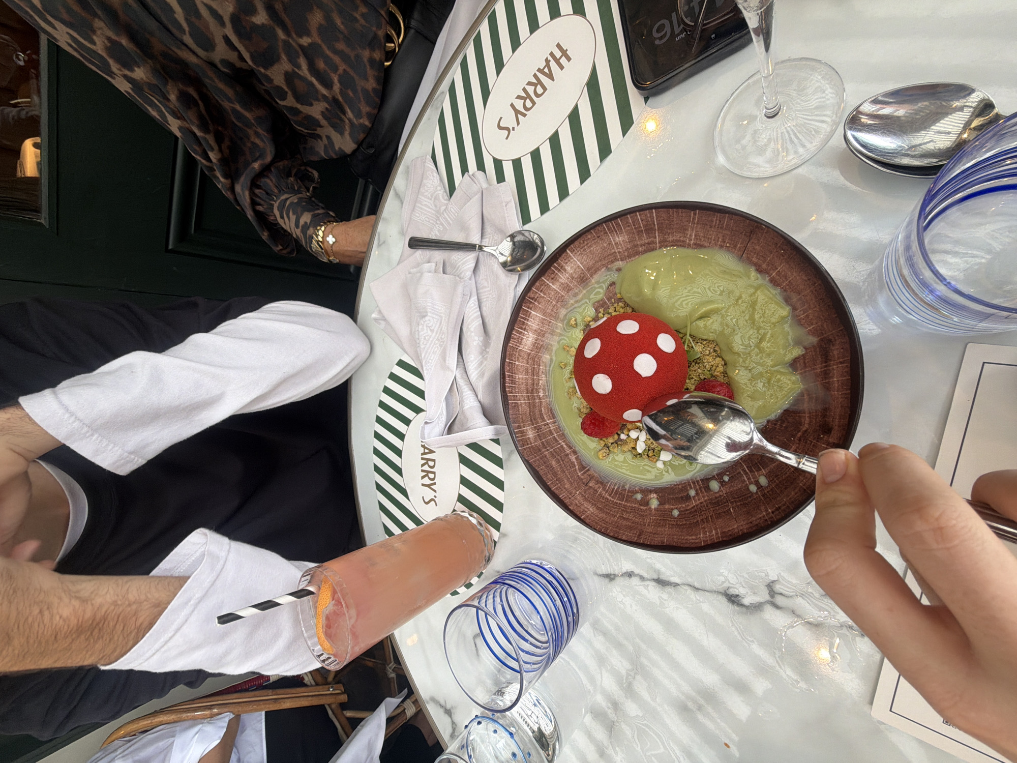 The image depicts a close-up overhead view of a table setting at a restaurant, likely in London, United Kingdom, as indicated by the "HARRY'S" branding on the placemats, suggesting a Harry's Bar location. In the foreground, a person's hand is visible, holding a spoon over a dessert. The dessert is presented in a brown, circular dish and features a vibrant green, semi-liquid base topped with what appears to be a red and white spotted mushroom-shaped confection, possibly a dessert mold, and some crumbled nuts or granola. A raspberry is also visible next to the mushroom.
To the left of the dessert, there is a tall glass containing a pink beverage, likely a cocktail, with a slice of citrus fruit as garnish and a black and white striped straw. Next to this drink, a shorter glass with blue stripes contains clear liquid. A white napkin with a subtle pattern is placed near the dessert, underneath a spoon. Another oval placemat with green and white stripes and the "HARRY'S" logo is partially visible.
In the background, parts of three individuals are visible. One person on the left is wearing a black long-sleeved shirt with white cuffs and collar. Another person in the center, partially obscured, appears to be leaning forward. To the right, a person is wearing a leopard-print long-sleeved garment, adorned with a bracelet featuring a clover charm. The table surface is a white marble-patterned material. A dark green doorway or wall is visible in the far background. A smartphone is also on the table to the right of the dessert. The overall scene suggests a leisurely meal or social gathering at a restaurant.