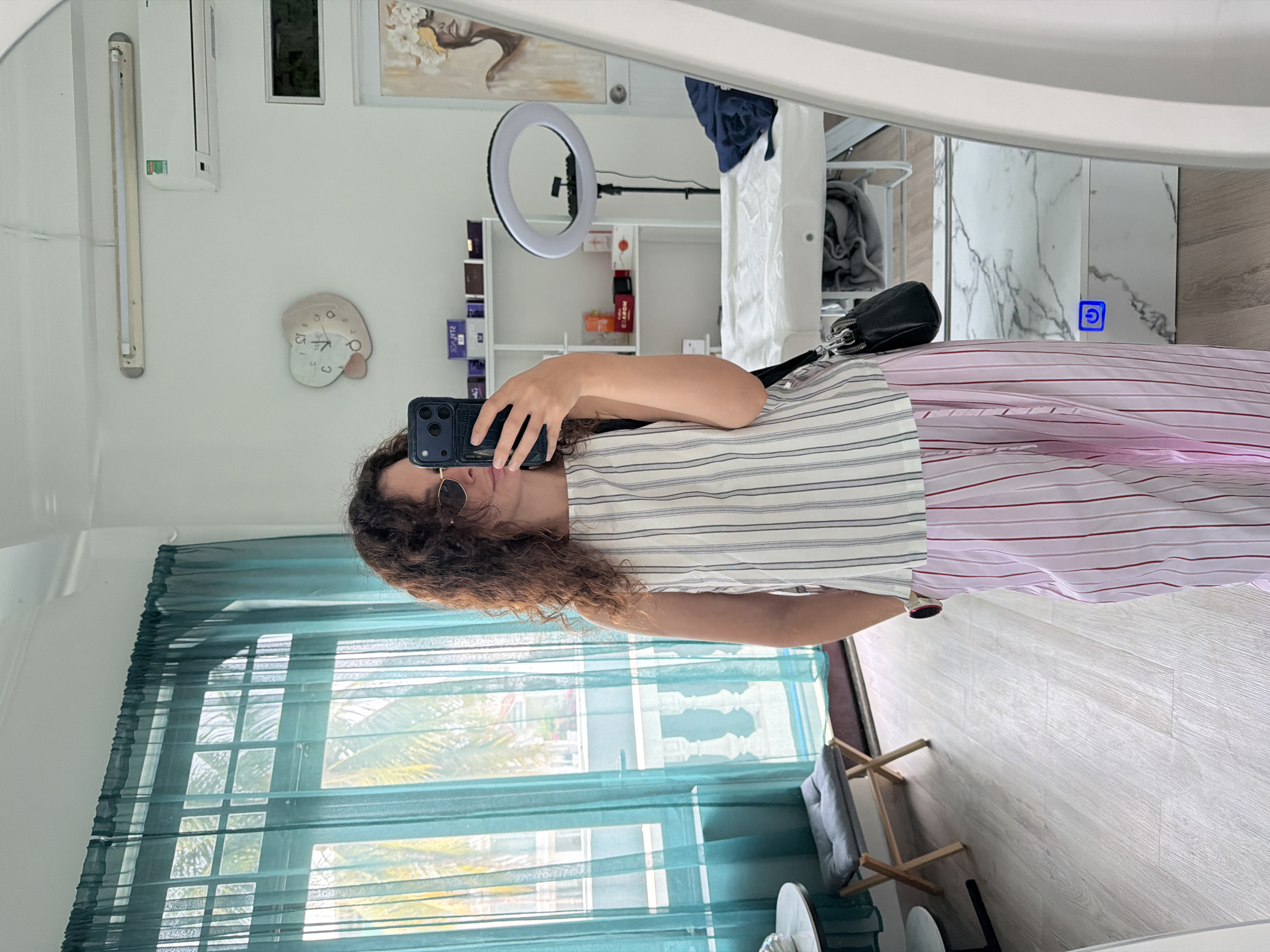 The image shows a young woman taking a mirror selfie in what appears to be a treatment room in the city of Bac Nha Trang Ward, Vietnam. She is wearing a striped sleeveless top and pink striped pants. She has curly brown hair and is wearing sunglasses. In the background, a treatment bed, shelves with products, a ring light, and a clock are visible. A teal curtain hangs by a window, and a folding stool with a gray cushion is placed near the window. The room has light-colored walls and wooden flooring. A blue power symbol is visible on a white surface in the mirror's reflection.