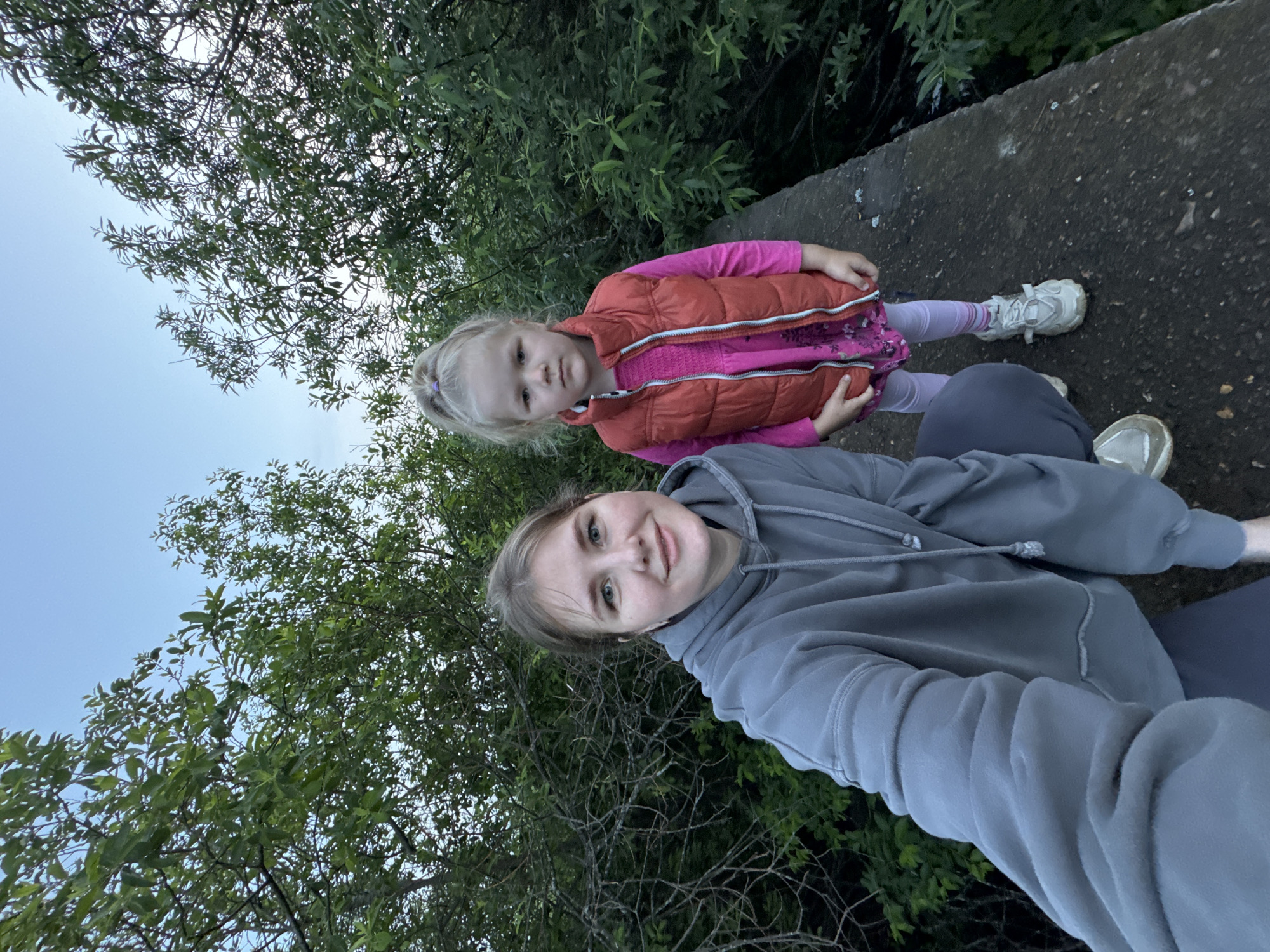 An outdoor, eye-level, full-shot selfie features two females in a wooded area. The woman on the left, in the foreground, is wearing a grey hooded sweatshirt and has her arm extended to take the photo. She has light skin, blue eyes, and light brown hair pulled back. She is smiling and looking at the camera. Next to her, to the right and slightly behind, is a young girl with blonde hair pulled into a high ponytail, wearing a pink long-sleeved shirt, a bright orange vest, light pink leggings, and white athletic shoes. She is looking directly at the camera with a neutral expression. They are standing on a concrete structure that appears to be a path or a bridge. The background is filled with dense green foliage and trees. The sky above is a pale blue, indicating either early morning or late evening light. The overall scene suggests a casual outdoor outing in a natural setting, likely near Горный, Russia, given the contextual information.