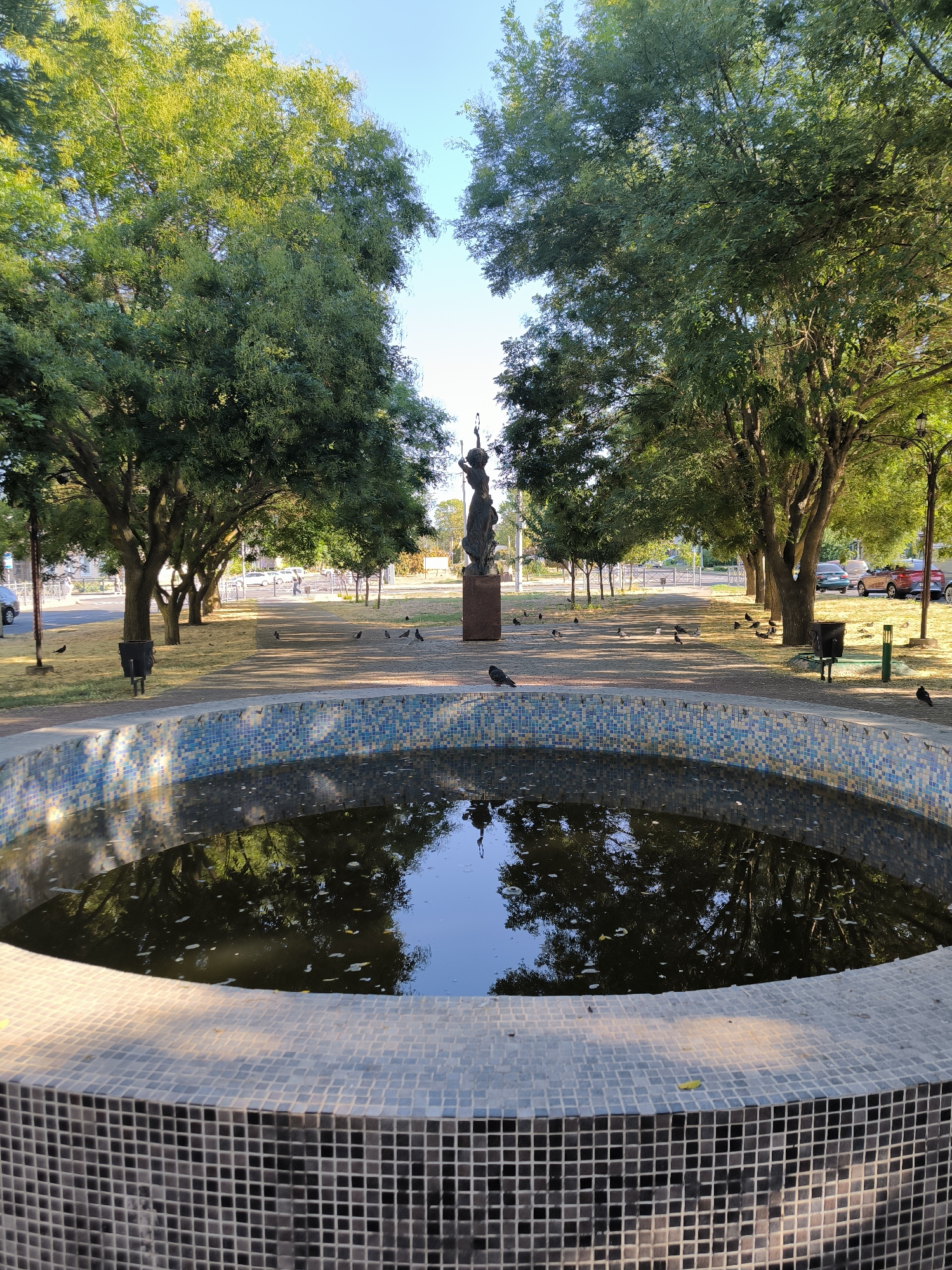 This image depicts a park with a central fountain in the foreground, which appears to be empty and reflects the surrounding trees and sky. The fountain is tiled with small blue, white, and grey mosaic squares. In the middle ground, a paved pathway is lined with trees on both sides, and a statue of a woman holding a torch stands prominently on a pedestal. Several pigeons are scattered throughout the pathway and the grassy areas. Beyond the pathway, a street is visible with parked cars and a few pedestrians. The environment suggests a sunny day in an urban park setting. The statue, a prominent feature in the park, is known as the Monument to the Founders of Mykolaiv and is located in the city of Mykolaiv, Ukraine.