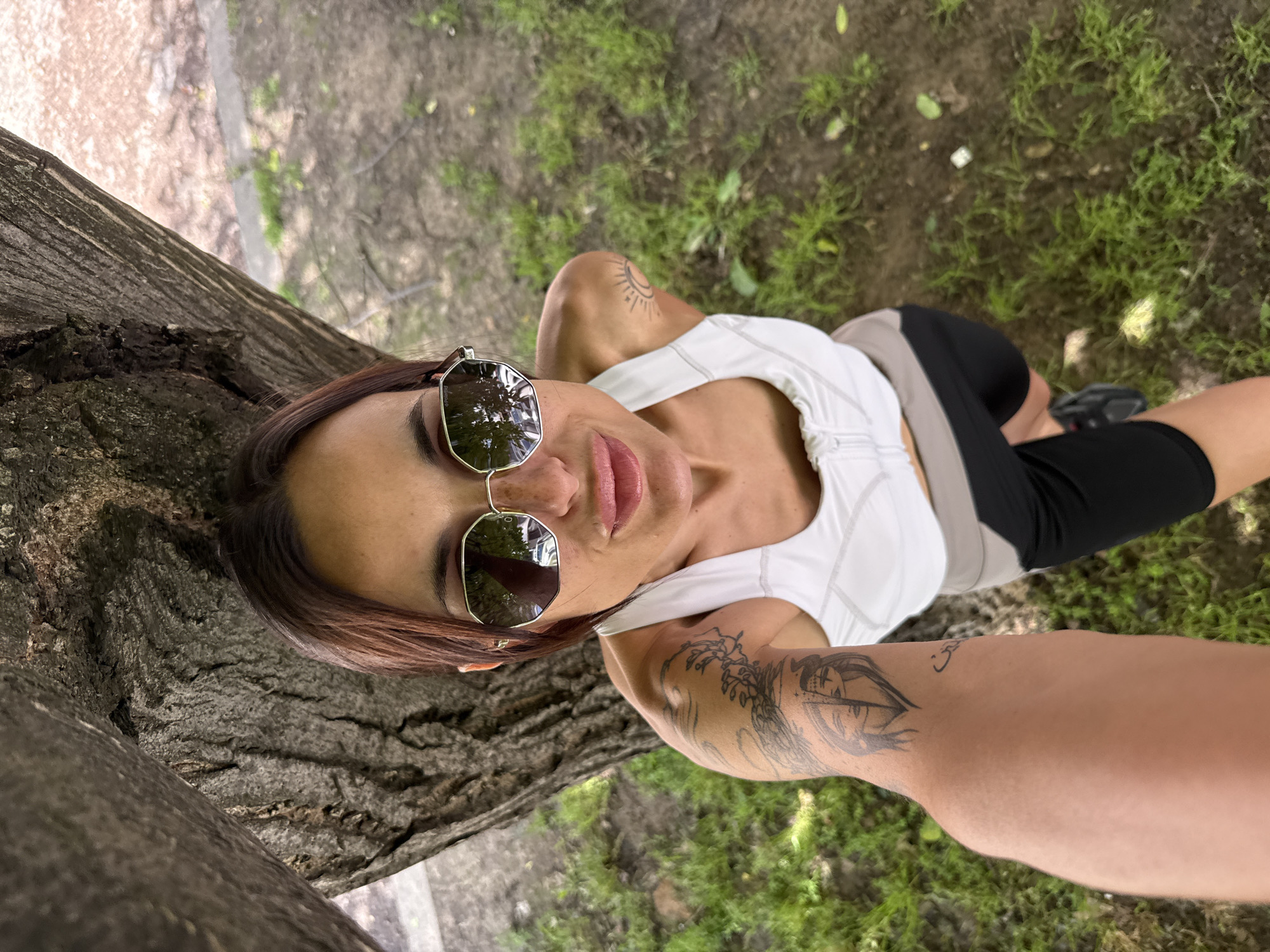 A woman is taking a selfie in a park. She is wearing sunglasses, a white tank top, and black exercise pants. She has tattoos on both arms. She is standing next to a tree, with grass and dirt visible in the background. The location could be a park in Pleven, Bulgaria.