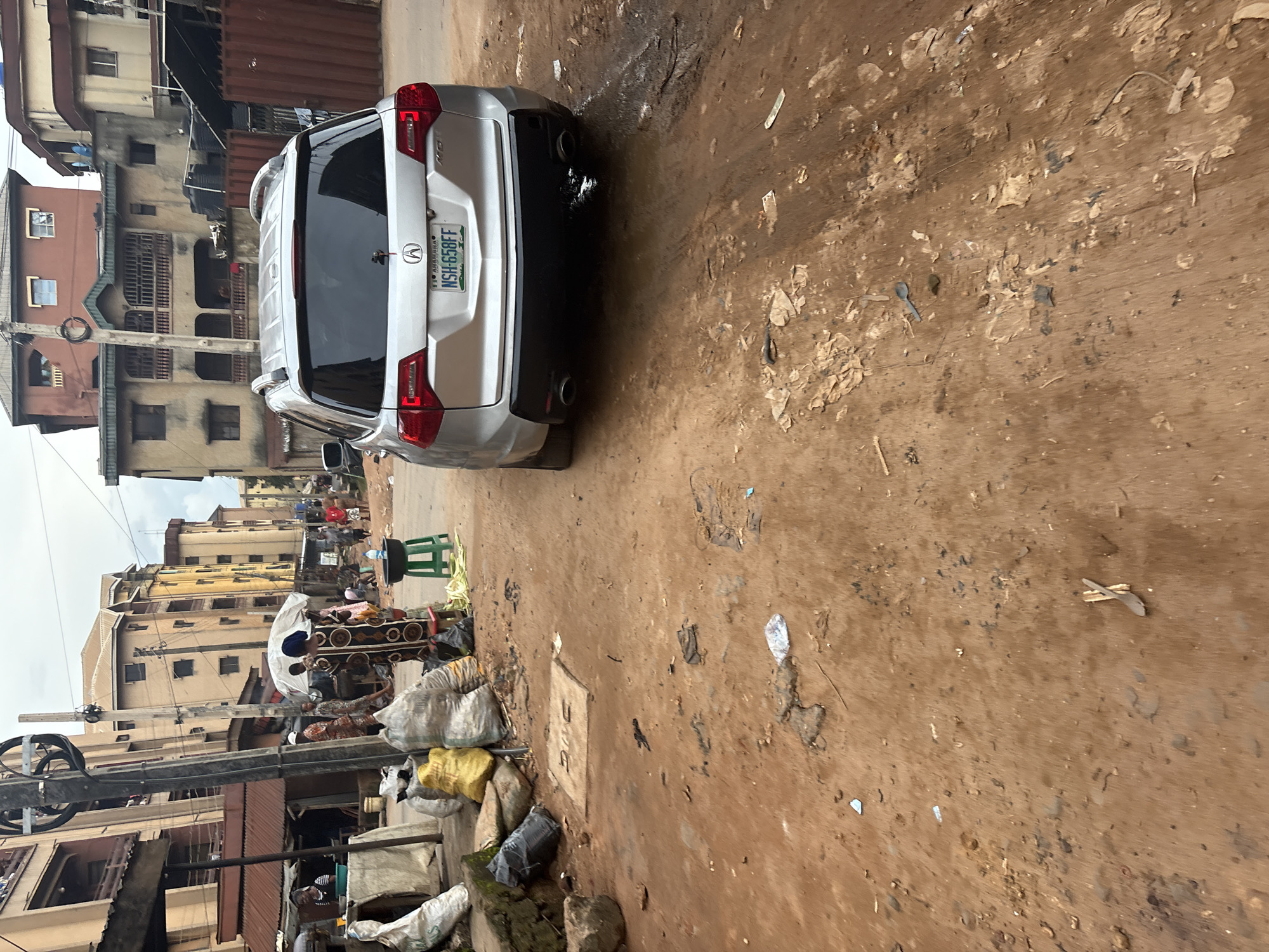 The image depicts a street scene in Onitsha, Nigeria, characterized by its urban environment. A silver Acura MDX SUV, with license plate N5H 658FF, is parked on a dirt road, its rear end facing the camera. The road is unpaved and covered with loose soil, debris, and some muddy patches, suggesting recent rain or poor drainage.
To the left of the vehicle, a busy street market is visible. Several individuals are engaged in commercial activities. A woman in a dark patterned dress stands near a white umbrella, possibly a vendor. Bags of goods, likely produce or other merchandise, are piled on the ground. Further down the street, other people are present, some walking and others gathered around stalls. The background shows multi-story buildings, typical of an urban African setting, with some exhibiting signs of wear. A utility pole with electrical wires stands prominently in the foreground. The overall atmosphere is that of a bustling, everyday scene in a Nigerian city, with a mix of residential and commercial spaces.