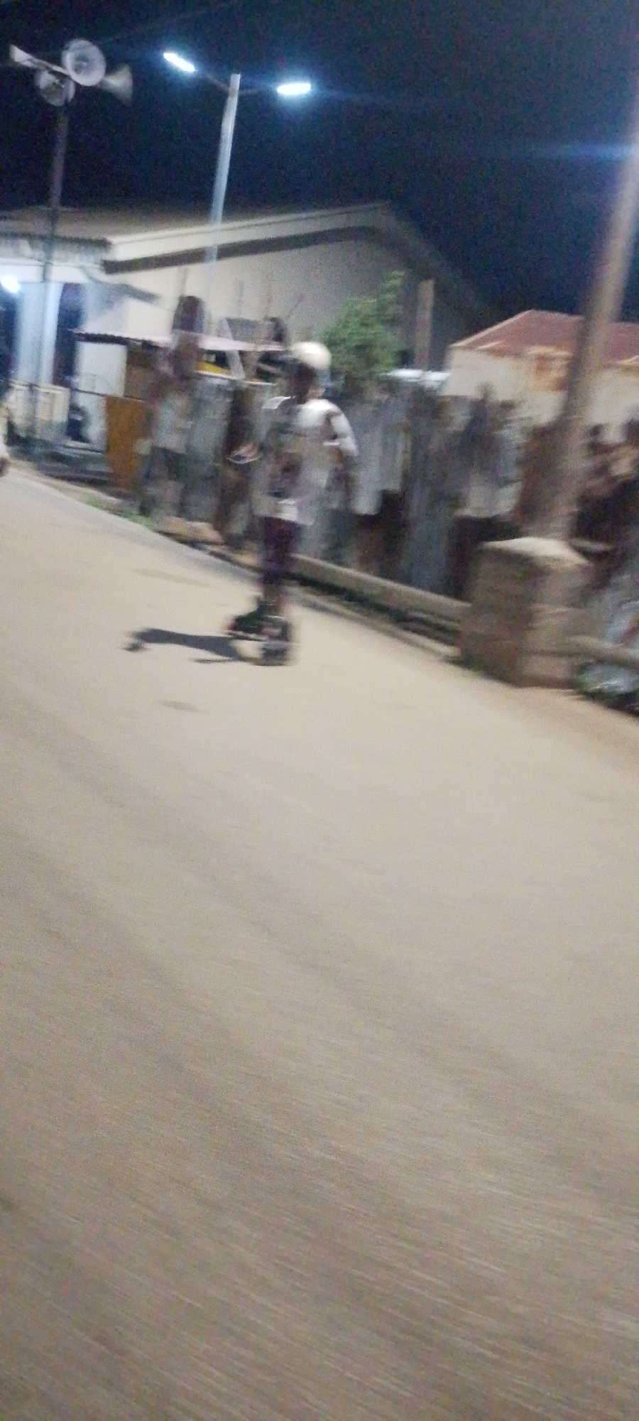 The image captures a nighttime scene in Jos, Nigeria, with a person on rollerblades in motion on a paved surface. The individual is wearing a light-colored shirt, dark pants, and a helmet. The surroundings are dimly lit, with streetlights visible in the background, illuminating buildings and vegetation. The motion blur indicates the photograph was taken with a moving camera or of a fast-moving subject, making definitive identification of specific landmarks or architectural styles difficult. The ground surface appears to be asphalt or concrete.