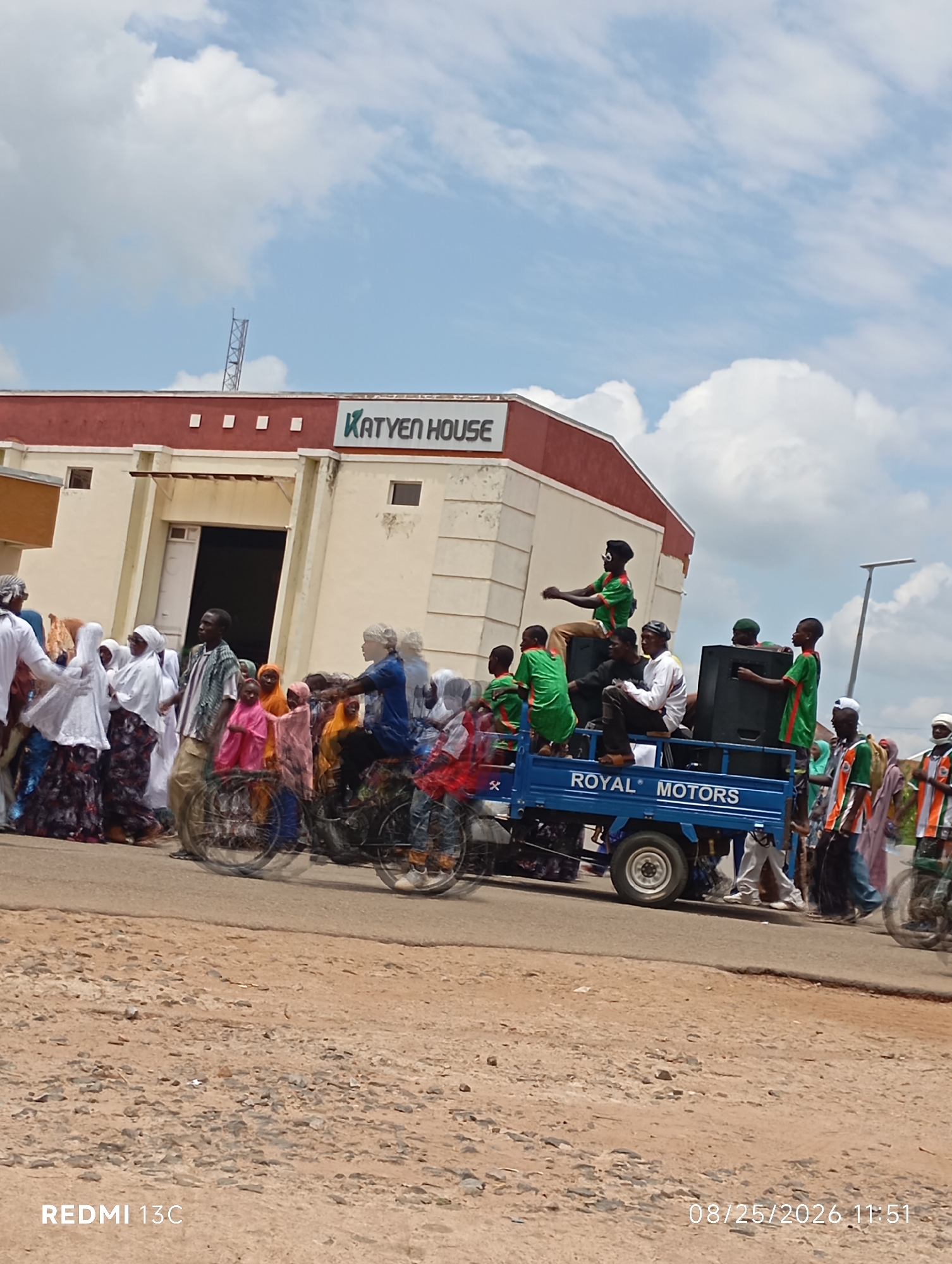 The image depicts an outdoor scene in Shendam, Nigeria. In the background, a building with "KATYEN HOUSE" signage is visible. In the foreground, a group of people are gathered, with some riding on a blue truck labeled "ROYAL MOTORS." Several individuals are dressed in green and orange attire. A few people are on motorcycles, and others are walking amongst the crowd. The environment appears to be a street with a dirt shoulder. The sky is cloudy. The timestamp at the bottom right indicates the image was captured on 08/25/2026 at 11:51. The bottom left corner shows "REDMI 13C".