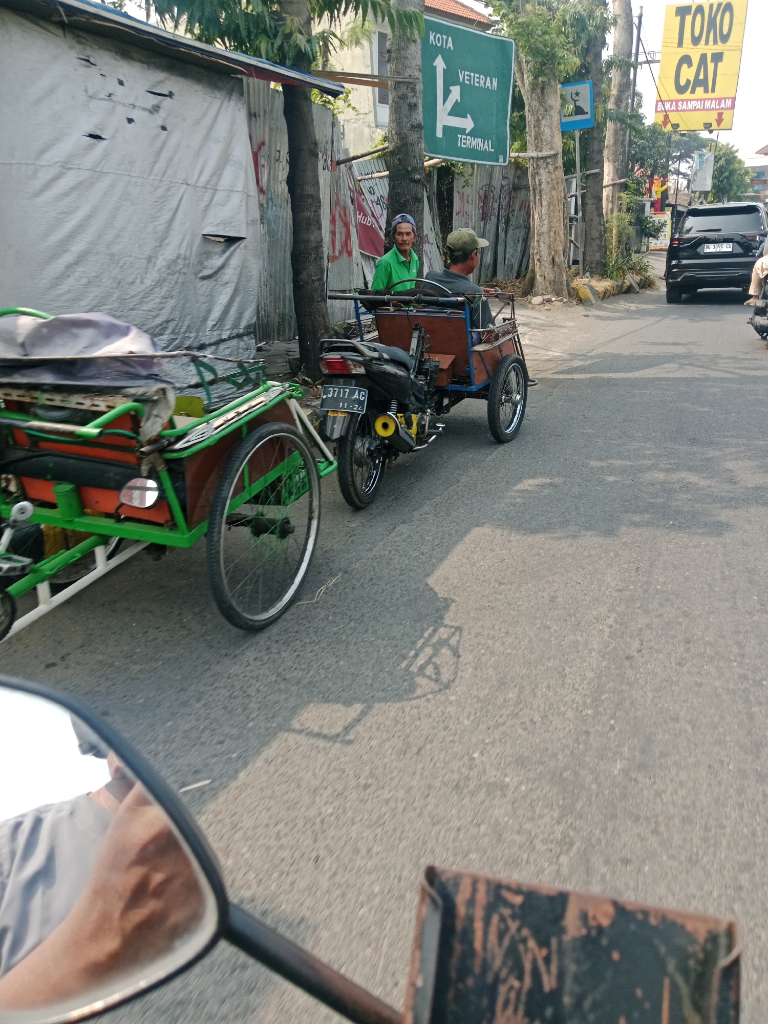 This image captures a street scene in Kediri City, Indonesia, on a bright, sunny day. In the foreground, a motorcycle-drawn cart with a green frame and an orange cargo box is parked on the asphalt road. The cart has a single rear wheel and a small front wheel. A black motorcycle is attached to the front of the cart, displaying a license plate that reads "3717 AG 11-24".
Two men are present in the scene. One man, wearing a green shirt and a blue cap, stands beside the cart, looking towards the camera. Another man, wearing a green cap, is seated on the cart, appearing to operate it. In the background, a black SUV is visible on the road, also with a license plate.
Various signs are posted along the roadside. A green sign indicates directions to "KOTA," "VETERAN," and "TERMINAL." A yellow sign reads "TOKO CAT" (Paint Shop) and "BUKA SAMPAI MALAM" (Open Until Night). Buildings with corrugated metal siding and some vegetation line the street. The overall impression is a typical urban street in Indonesia, bustling with everyday activity.