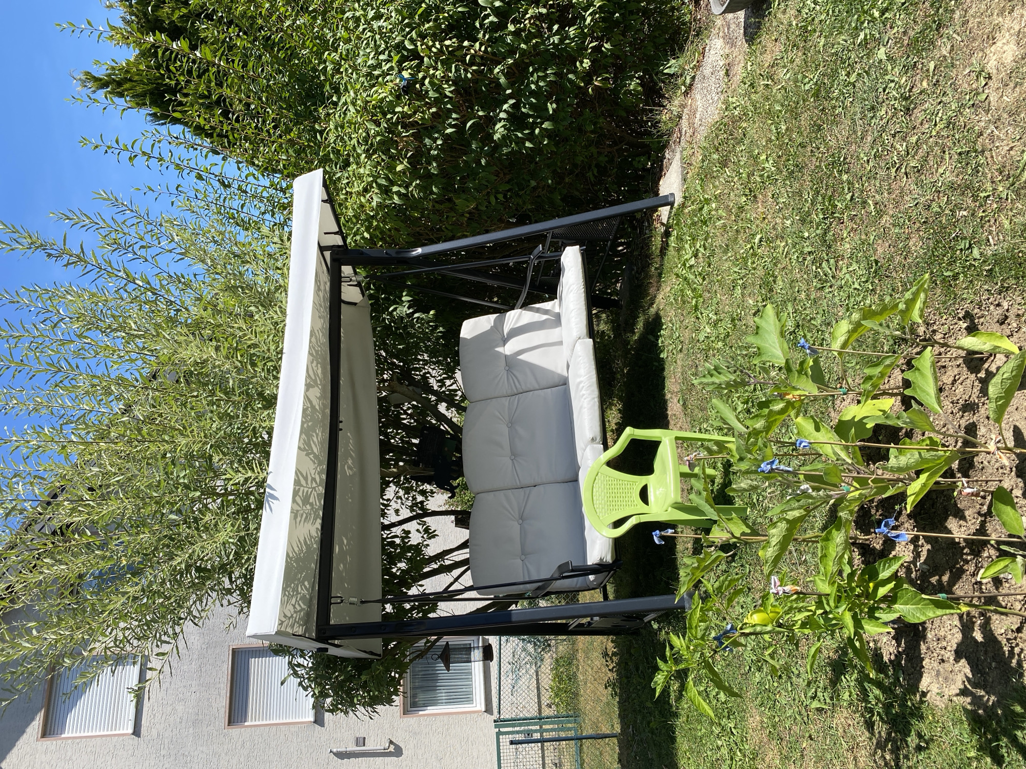 A sunny, exterior shot captures a backyard swing set with a white canopy and three white cushions. A small, bright green plastic chair sits in front of the swing set, facing the camera. In the foreground, several small plants with wooden stakes and blue ties are visible in a patch of dirt. The background features a building with light-colored textured walls and windows with white shutters, as well as lush green trees and bushes. The scene is set in Flammersfeld, Germany.
