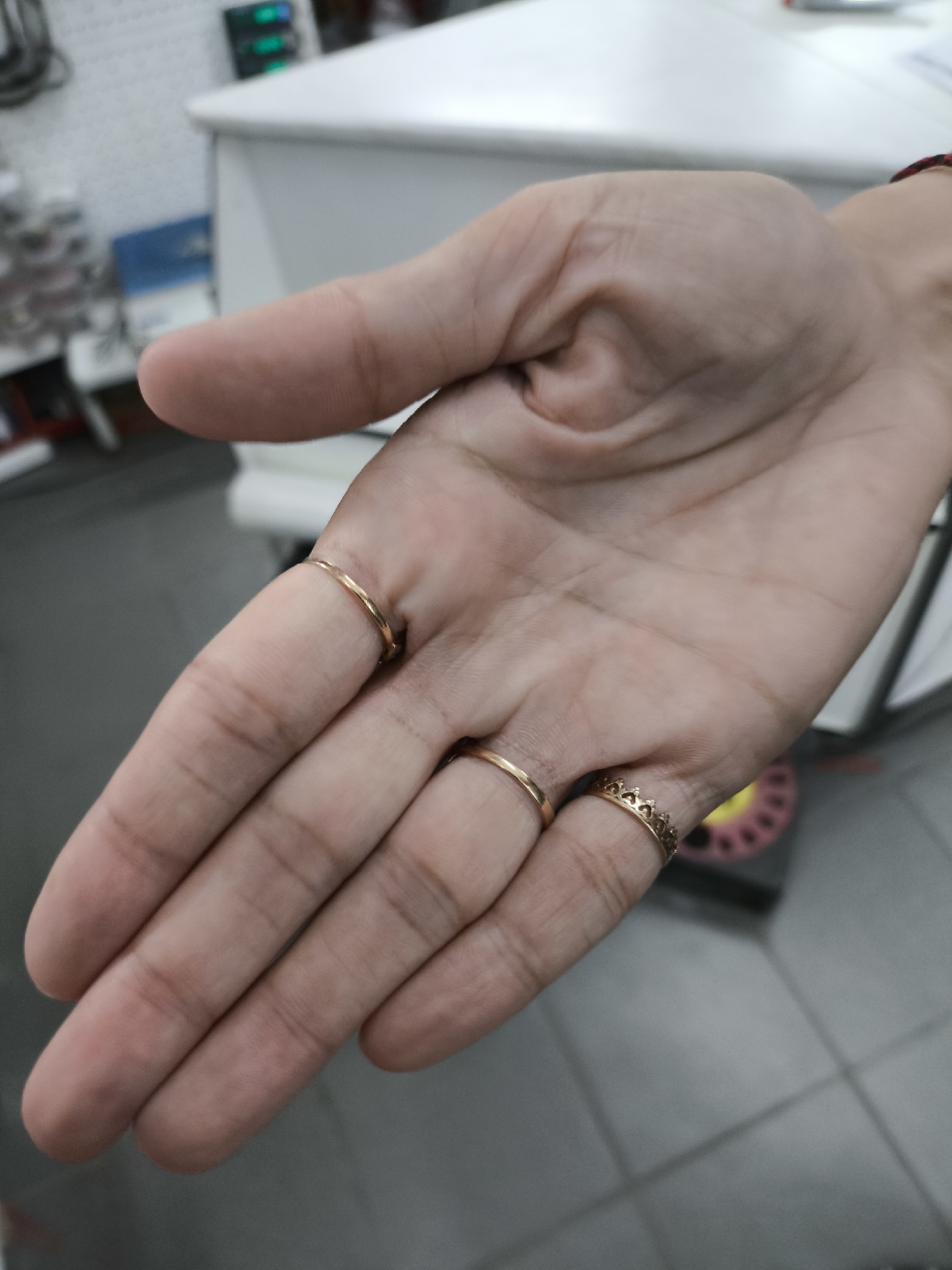 The image shows a close-up of a person's left hand, palm up, with three rings. One simple gold band is on the ring finger, a second gold band is on the middle finger, and a gold ring shaped like a crown is on the index finger. The background is blurred, showing what appears to be an indoor setting with a white counter and tiled flooring. There is no information in the image to suggest the location is Solikamsk, Russia.