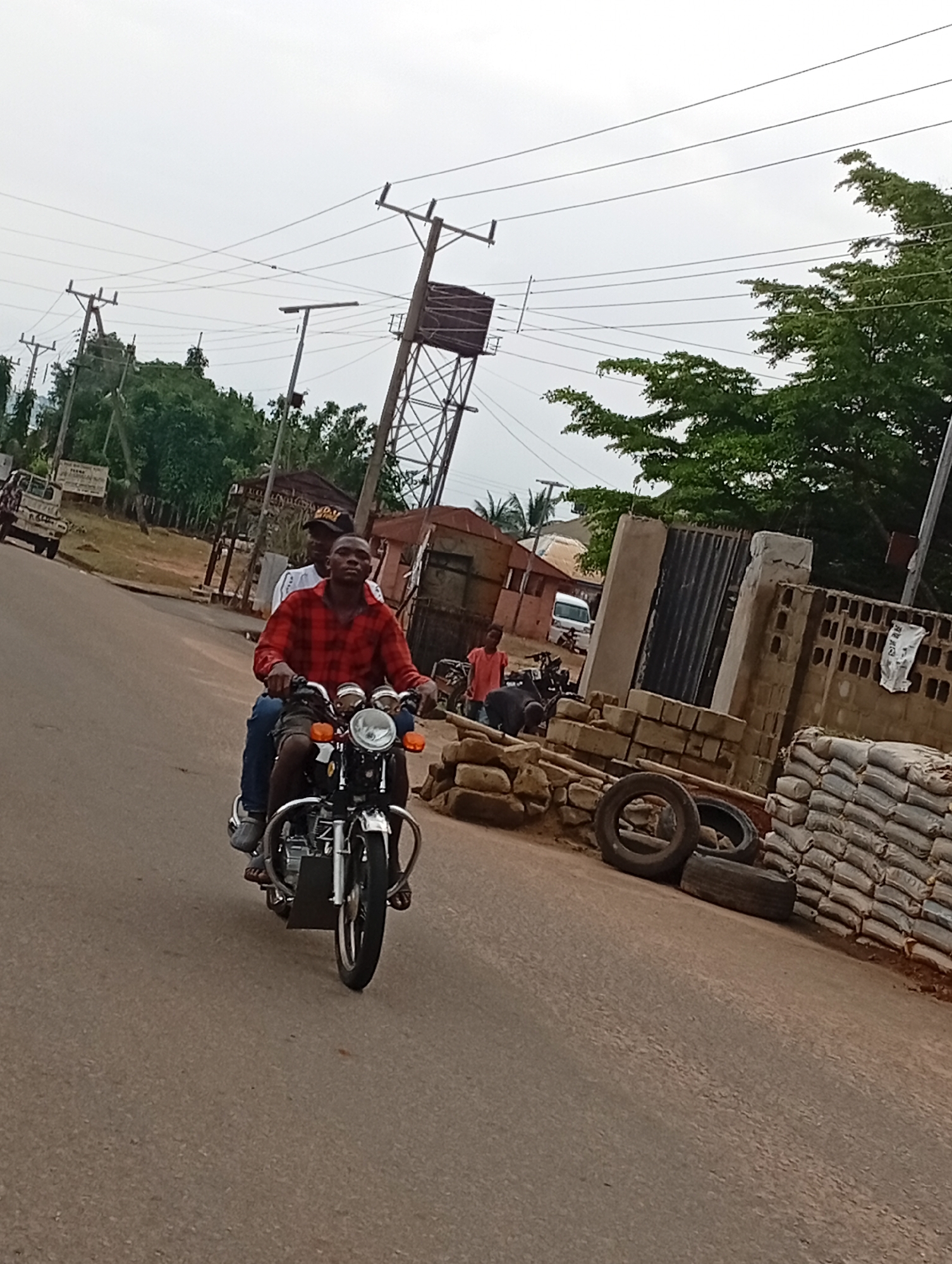 This is a medium-shot, eye-level, outdoor, daytime photograph taken on an overcast day in Nsugpe, Nigeria. The scene depicts a street with a two-person motorcycle in the foreground, traveling from left to right. The driver, a man wearing a red plaid shirt and dark pants, is in the front, looking forward. Behind him, a second man, wearing a white shirt and a dark cap, is seated on the motorcycle. The motorcycle is a standard model with a chrome fender and handlebars, and orange turn signals.
The street is paved with asphalt. To the right of the road, there is a wall made of cinder blocks and stones, with stacks of white bags, possibly cement, piled against it. Several tires are also visible on the ground near the wall. Behind the wall, there are buildings with corrugated metal roofs and a tall metal water tower.
To the left of the road, there are more buildings, trees, and utility poles with power lines crisscrossing overhead. A white truck is parked on the side of the road in the background. The overall environment suggests a bustling, somewhat rural or semi-urban area.