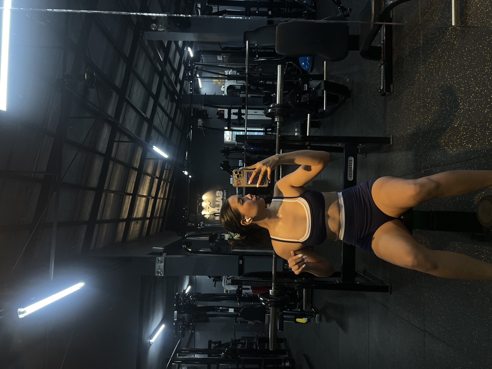 A young Asian woman with dark hair tied back in a ponytail is taking a mirror selfie in a gym. She is wearing a dark blue sports bra with a white trim and matching dark blue shorts with a white waistband. She is holding a smartphone in her right hand, angled to capture her reflection. The gym appears to be well-equipped with various weight machines and benches visible in the background. The lighting is provided by fluorescent tubes on the ceiling. The image is taken from a medium distance, showing the woman from approximately the knees up, with the gym environment filling the rest of the frame. The gym is located in Tanza, Philippines.