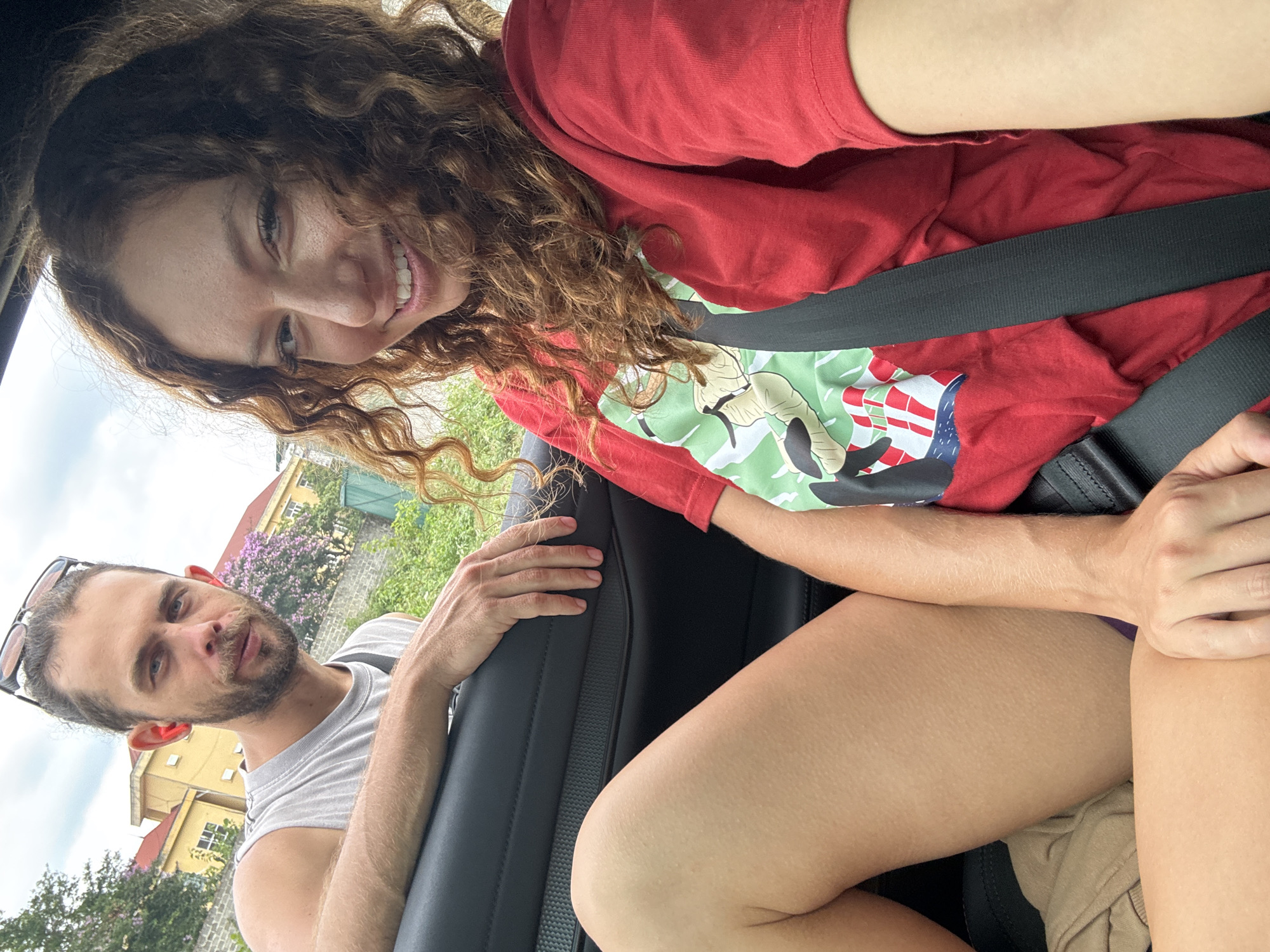 From a low-angle perspective inside a car, a woman with curly brown hair and a red t-shirt is smiling while wearing a black seatbelt. A man with a trimmed beard and sunglasses atop his head is in the background to the left, with his arm resting on the car door. Outside the car are buildings and greenery. Based on architectural details and foliage, the environment suggests a street scene in Trung Tam Ward, Vietnam.