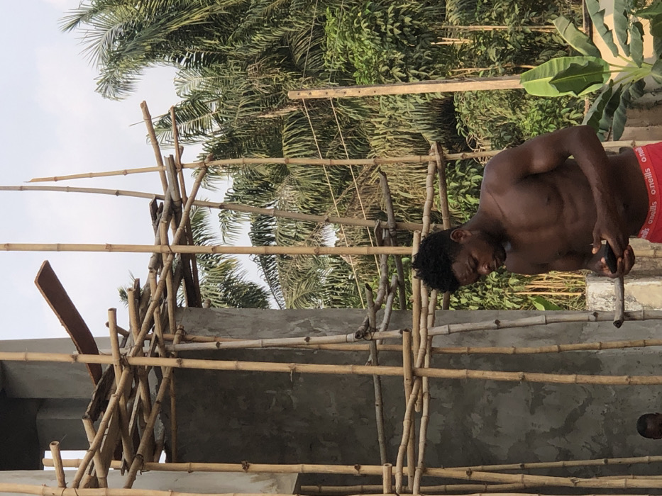 An adult male stands shirtless, facing forward and looking down at a black smartphone held in both hands. He has a dark complexion, a muscular build, and short dark curly hair. He wears red boxer briefs with "O'NEILLS" branding. To his left, a concrete building structure is under construction, surrounded by extensive bamboo scaffolding secured with lashings; wooden planks are positioned across parts of the scaffolding. A small child's head is partially visible in the lower-left corner, obscured by the concrete. In the background, dense tropical vegetation, including numerous palm trees and a banana plant, is visible under a bright sky. A wooden utility pole with visible wires stands on the right. The scene depicts construction activity within a vegetated area in Enugwu-Ukwu, Nigeria.