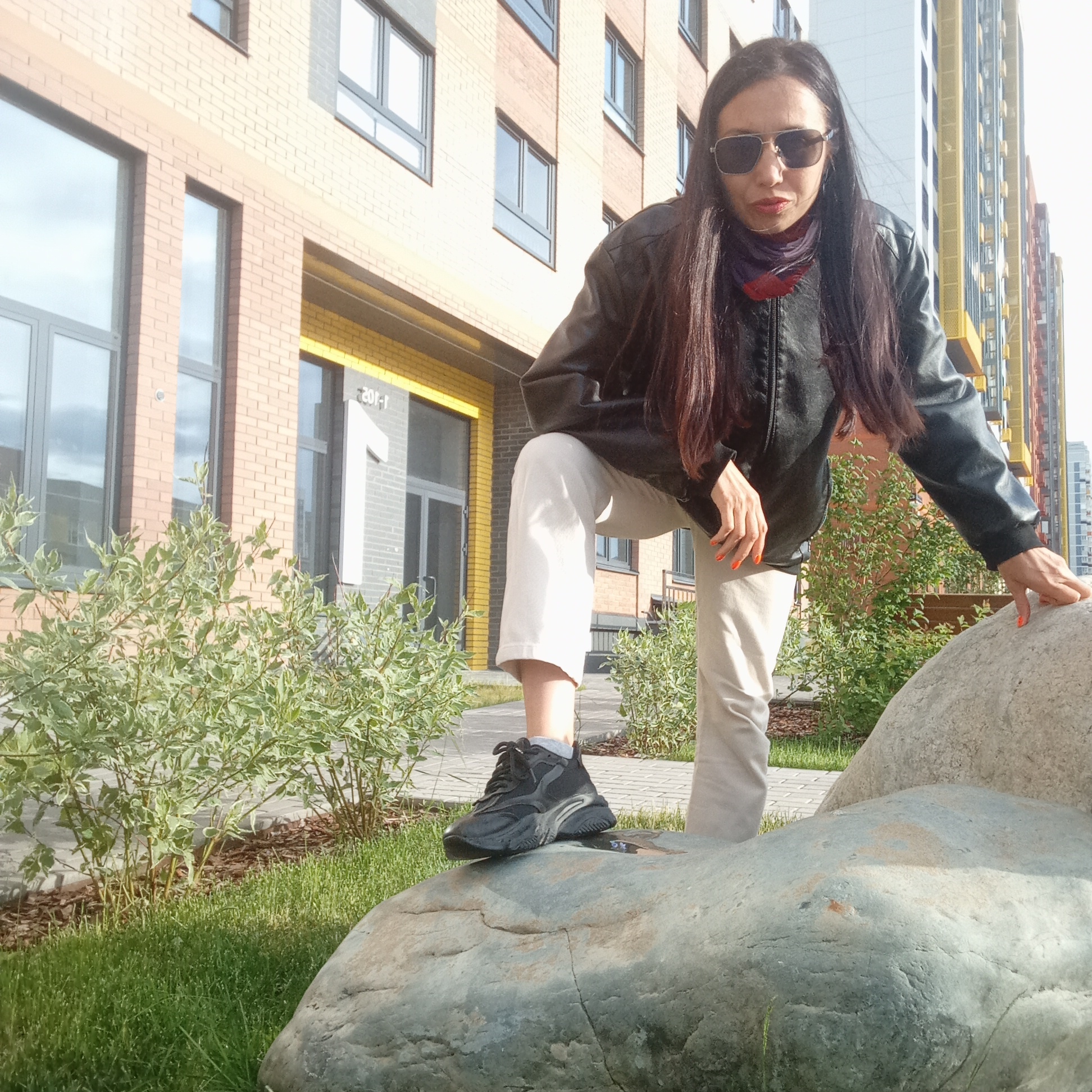 This outdoor full-body shot features a woman posing confidently in front of modern residential buildings in Izhevsk, Russia. She stands with her right foot placed firmly on a large, smooth, grey-blue rock, while her left foot is on the ground. She leans forward slightly, with her right hand resting on her knee and her left hand touching another rock. She looks directly at the camera through dark aviator-style sunglasses.
The woman has long, dark hair and is dressed in a black leather jacket, a multi-colored scarf (appearing in shades of purple and red), light beige or off-white cropped pants, and black athletic sneakers. Her fingernails are painted a bright orange.
The immediate surroundings include multiple large rocks, lush green grass, and small green bushes with variegated leaves, indicating a landscaped area. Behind her, a paved walkway is visible. The background consists of modern, multi-story apartment buildings. These buildings feature facades of light brown and grey brick, with numerous rectangular windows. One building to the far right also showcases striking yellow and blue vertical paneling. On the ground floor of a building behind the woman, the number "1" is prominently displayed, along with the text "201-1" near an entrance. The bright, clear sky and strong sunlight suggest a sunny day.