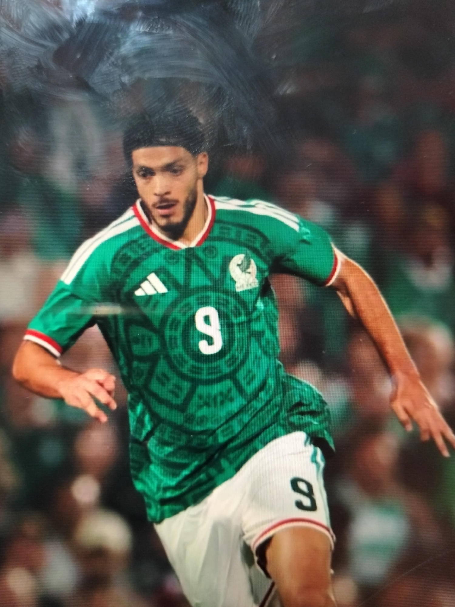 A male soccer player, wearing a green jersey with white trim and the number 9 on the front and shorts, is shown in profile, facing left. The jersey features a circular Aztec-inspired design and the Adidas logo. The player has dark, curly hair and a beard, and appears to be in motion. The background is blurred, suggesting a stadium setting with an audience. The image is a photograph, and it is not possible to determine the specific location in the city of Payagaboh, Indonesia.