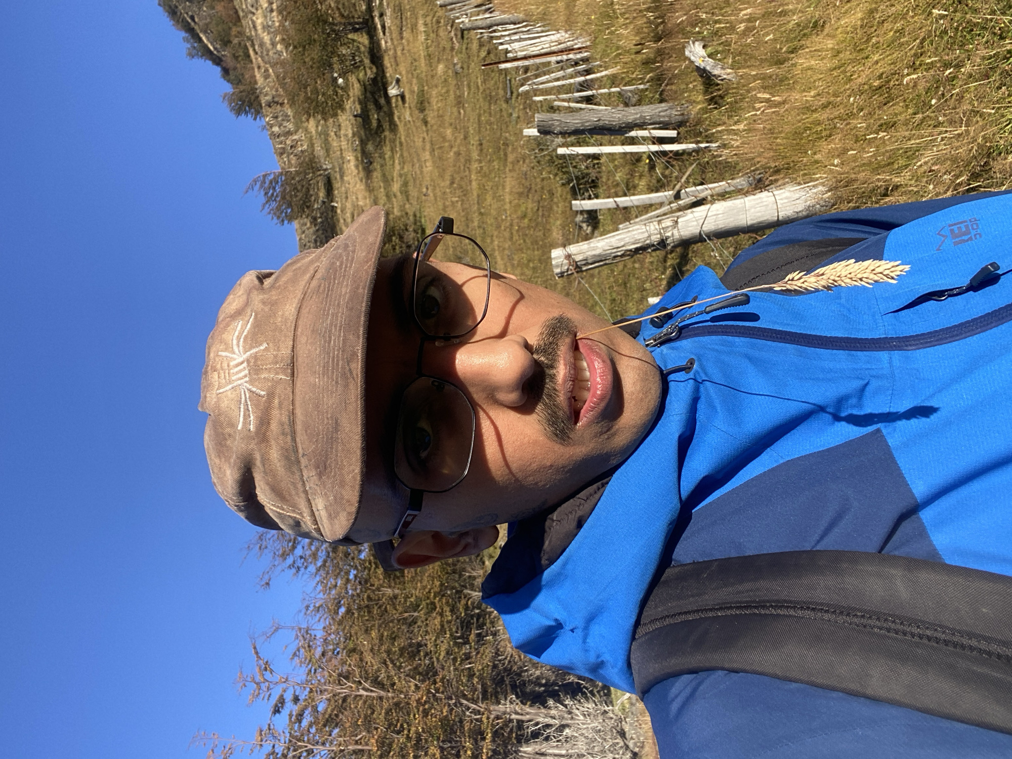 A person wearing a brown cap, glasses, and a blue jacket with a backpack is positioned in front of a rural landscape. The individual is facing the camera with a piece of wheat between their lips. The background shows a field with dried grass and a decaying wooden fence running along the field's edge. Bare trees are interspersed throughout the landscape. The sky is clear and blue. The person is in Ushuaia, Argentina.