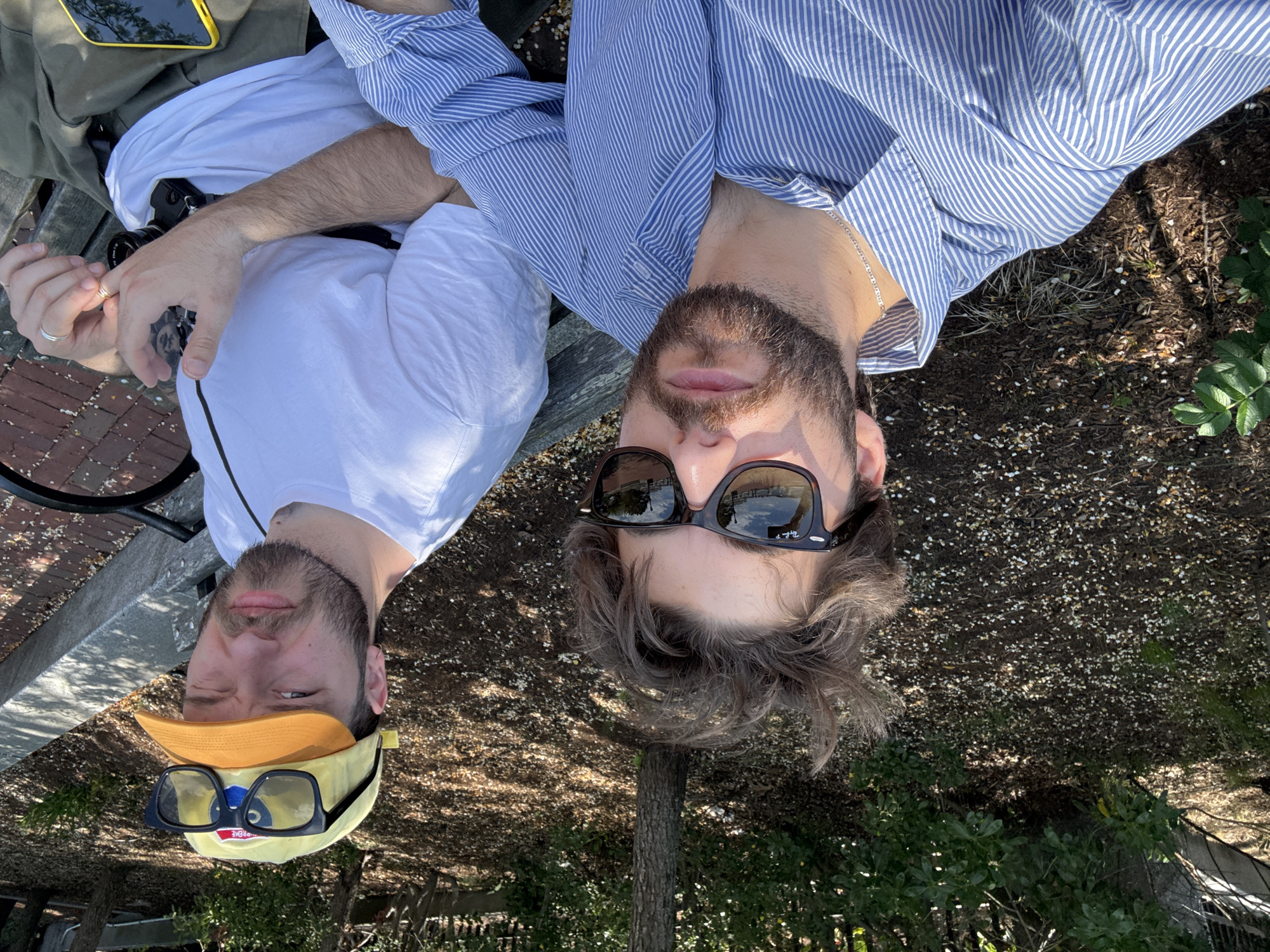 Two men are reclining on a bench outdoors. One man wears a blue and white striped shirt and dark sunglasses. The other man wears a white shirt and a yellow baseball cap with dark-framed glasses on top of it. The location is outdoors with a view of trees and a fence.