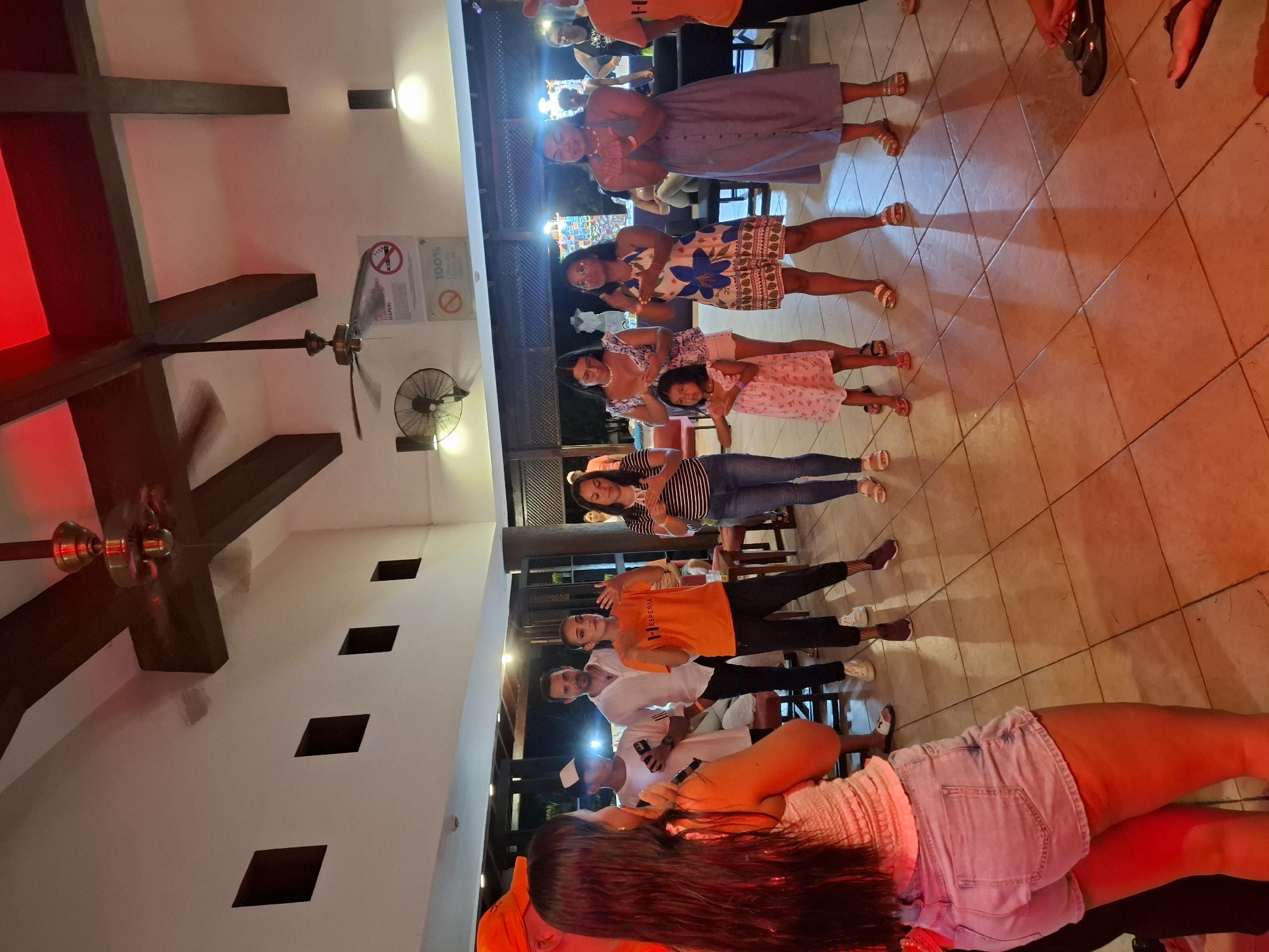 A group of people is photographed in a lighted area that is possibly a restaurant or bar in Playa El Agua, Venezuela. A group of seven people is seen posing and facing forward, while another person is shown in the foreground lying down on a chair or bench with her back facing the camera. An indoor ceiling fan is visible, as well as beams and what appear to be lighting fixtures. A sign with symbols of a smoking ban and 100% cleanliness is mounted on the wall.