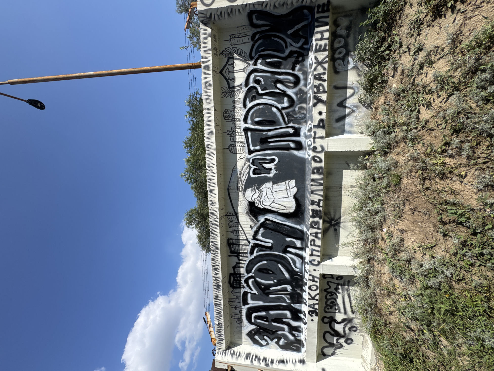 A graffiti-covered concrete barrier on a sunny day in Oskemen, Kazakhstan, features stylized lettering in black and white spray paint. The large lettering reads "ЗАКОН И ПОРЯДОК" (Law and Order). Below this, smaller text says "• ЗАКОН • СПРАВЕДЛИВОСТЬ • УВАЖЕНИЕ •" (Law, Justice, Respect). A drawing of a person in a hooded cloak is depicted between the large lettering, and a stylized drawing of a courthouse with scales of justice is located to the right of the lettering. The barrier is situated on a sloped embankment covered with green vegetation. A rusty lamppost stands to the right of the barrier against a blue sky with white clouds.