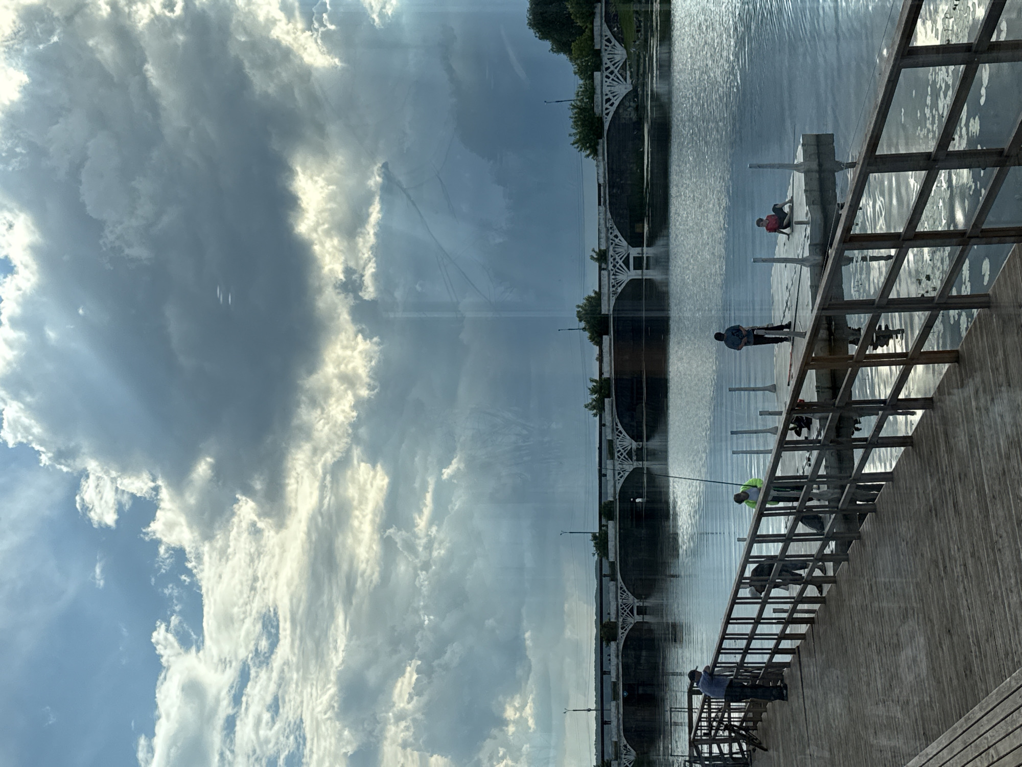 The image shows a body of water with a bridge in the background, reflecting the sky and clouds. The sky is partly cloudy with bright white cumulus clouds and some darker, grayer areas, suggesting a dynamic weather pattern. The water's surface is rippled, indicating a gentle breeze.
In the foreground, there is a wooden walkway or pier with a metal railing. Several people are visible on the walkway, some fishing and others appearing to be working. One person in a bright green vest is on a ladder that extends over the water, possibly engaged in some kind of maintenance or construction. Another person in a red shirt is sitting on the walkway. The scene suggests a daytime activity, possibly late afternoon given the angle of the light.
The bridge in the background is an arched structure with decorative white railings, lined with trees. The overall mood is calm and observant, with the focus on the interplay between the natural elements of sky and water and the human activities taking place. The image is taken from a slightly elevated, angled perspective, looking across the water towards the bridge. There is no visible text in the image.