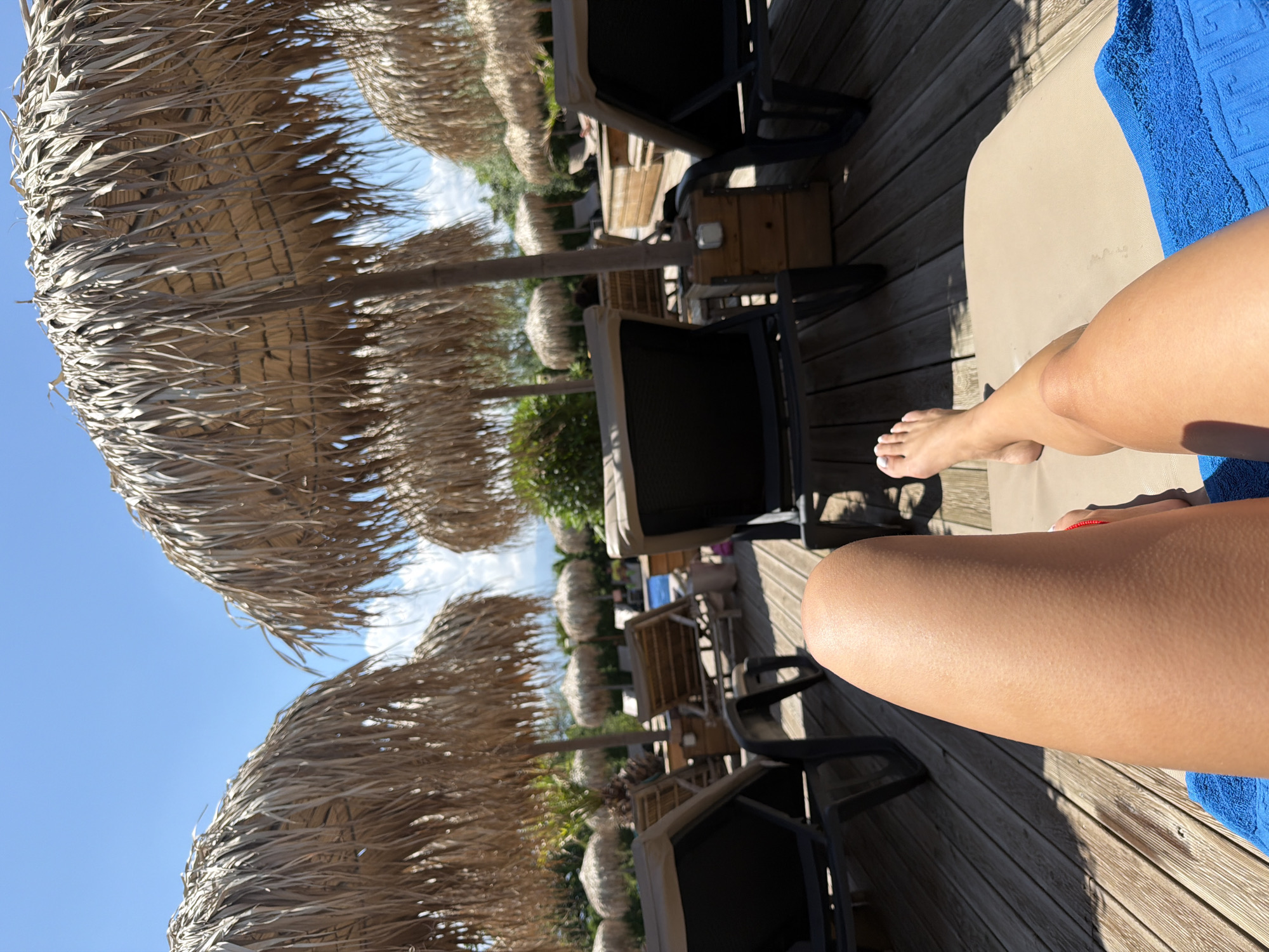 A person's legs are positioned on a lounge chair by a swimming pool, with a blue towel underneath. The foreground shows the person's tanned legs, with one foot visible at the top right. In the background, there are several thatched umbrellas providing shade over lounge chairs. The setting appears to be a resort or a private outdoor area, with wooden decking and lush greenery. The sky is clear blue with a few white clouds. The location is not explicitly identifiable as being in Plovdiv, Bulgaria.