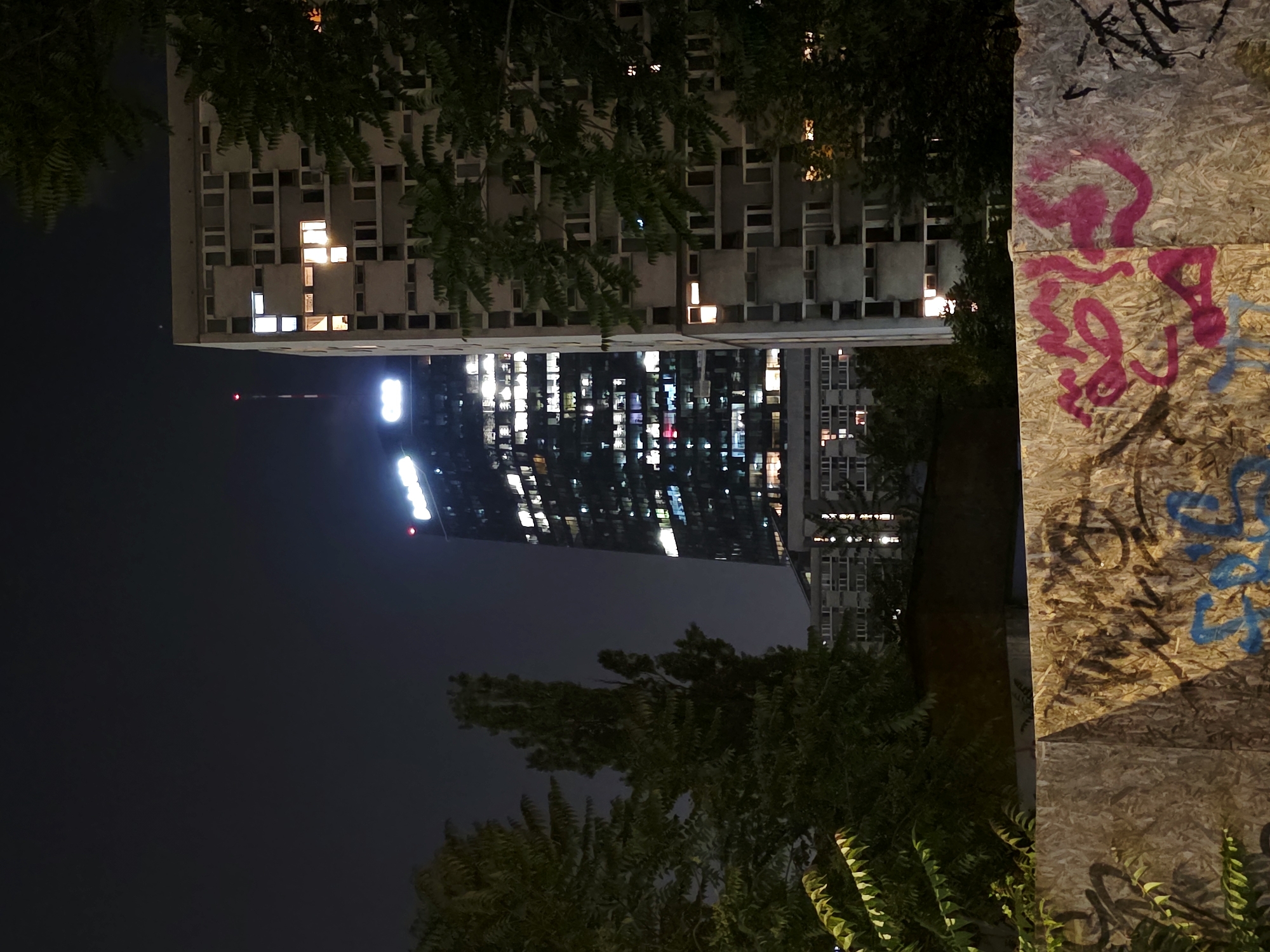 The image captures a nighttime urban scene in Warsaw, Poland, likely in the vicinity of the Rondo ONZ area. In the background, the prominent Złota 44 skyscraper, a modern residential high-rise, is illuminated, its numerous windows glowing with artificial light. To its right stands a Brutalist-style residential building, also partially lit, with a repeating pattern of windows and balconies. Trees with dark foliage are visible in the mid-ground, obscuring parts of the buildings and the sky. In the foreground, a low wall constructed of plywood is covered in graffiti, with vibrant pink and blue spray paint tags visible. The overall impression is one of a city at night, with a mix of modern and older architecture, contrasted by street art in the foreground. The sky is dark and overcast.