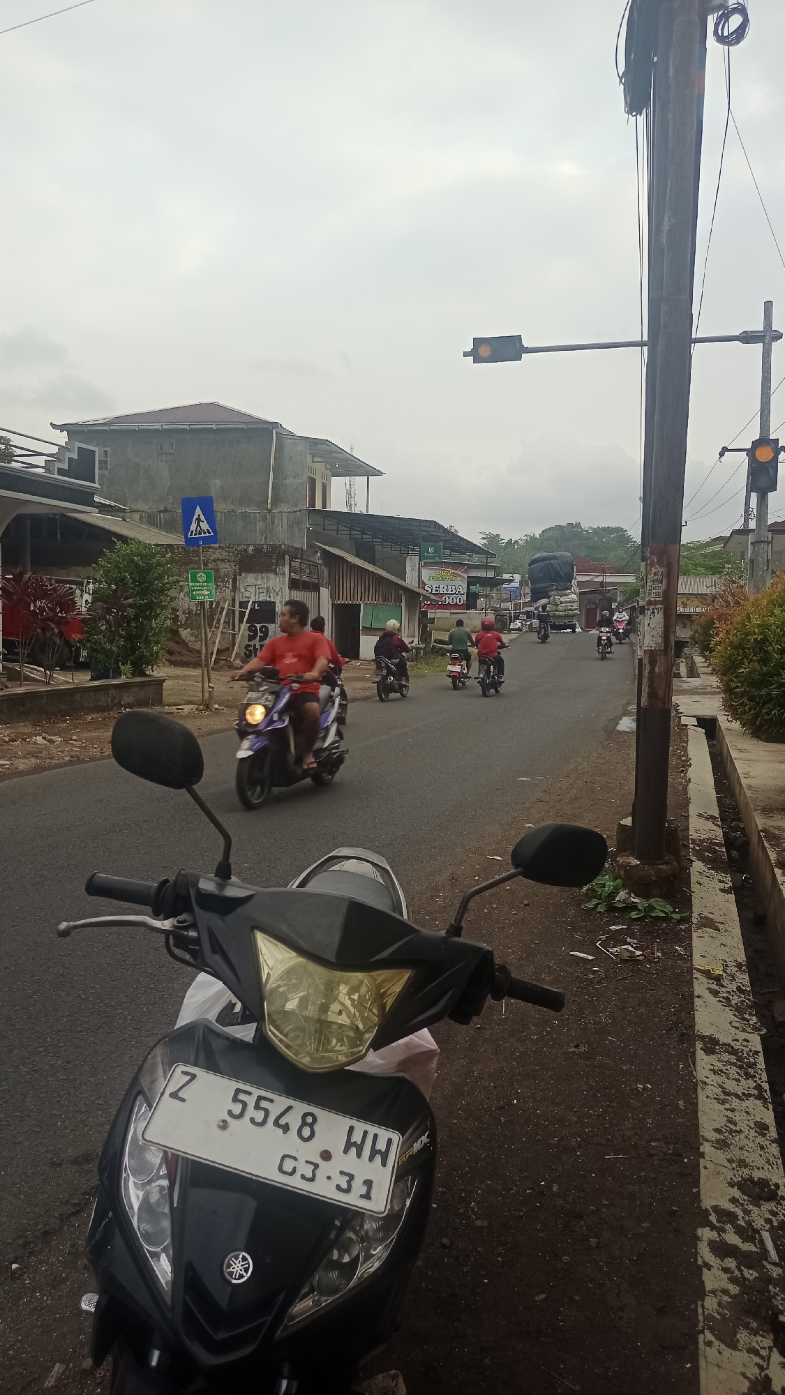 A street scene in Karyamandala, Indonesia, under a cloudy sky. In the foreground, the front of a black Yamaha motorcycle is visible, with its license plate displaying "Z 5548 WH C3.31". The motorcycle is parked on the side of a paved road. Several other motorcycles are traveling on the road, with riders wearing casual clothing. One rider in a red shirt is prominent in the mid-ground. Buildings line the left side of the road, appearing to be shops or residences. A traffic light, showing yellow, is visible hanging over the road. A utility pole with wires is positioned to the right of the road. The environment is a typical urban or semi-urban setting with moderate vegetation and signs of daily life.