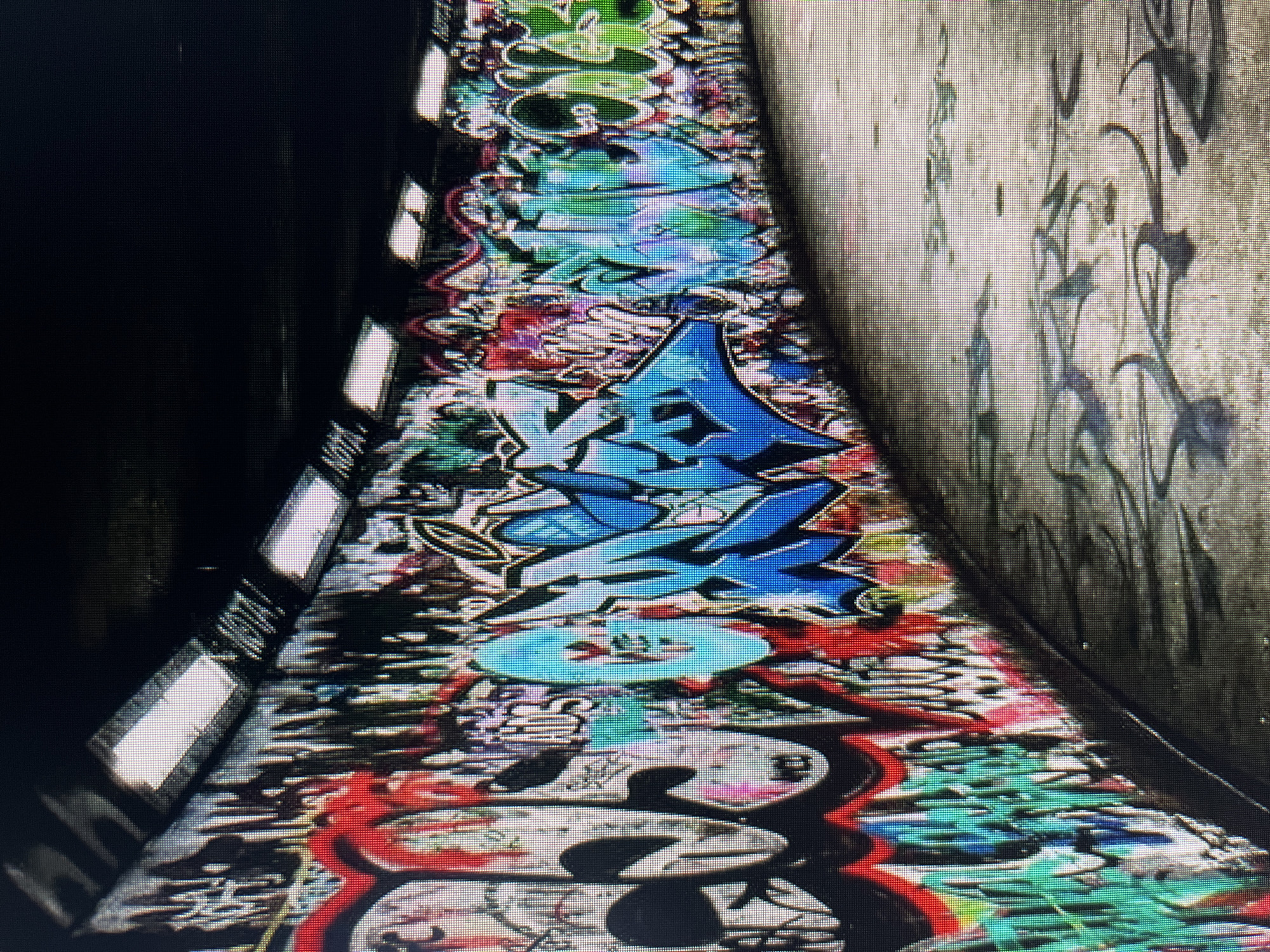 The image shows a skatepark with walls covered in vibrant graffiti. The skatepark is an underground or covered structure with a curved wall on the left and a flat, concrete surface on the right. The wall is densely covered in graffiti in various colors and styles, including bold lettering, abstract designs, and cartoon-like figures. The graffiti ranges from red, black, and white to bright blues, greens, and purples. The ceiling of the structure has several fluorescent lights, which illuminate the area. The ground of the skatepark is concrete and shows signs of wear and tear, with some graffiti or paint marks visible on it as well. There are no people or animals visible in the image. The overall impression is one of urban art and recreation in a somewhat gritty, industrial setting. The location is described as Unknown, Unknown, indicating that specific geographical information is not available.