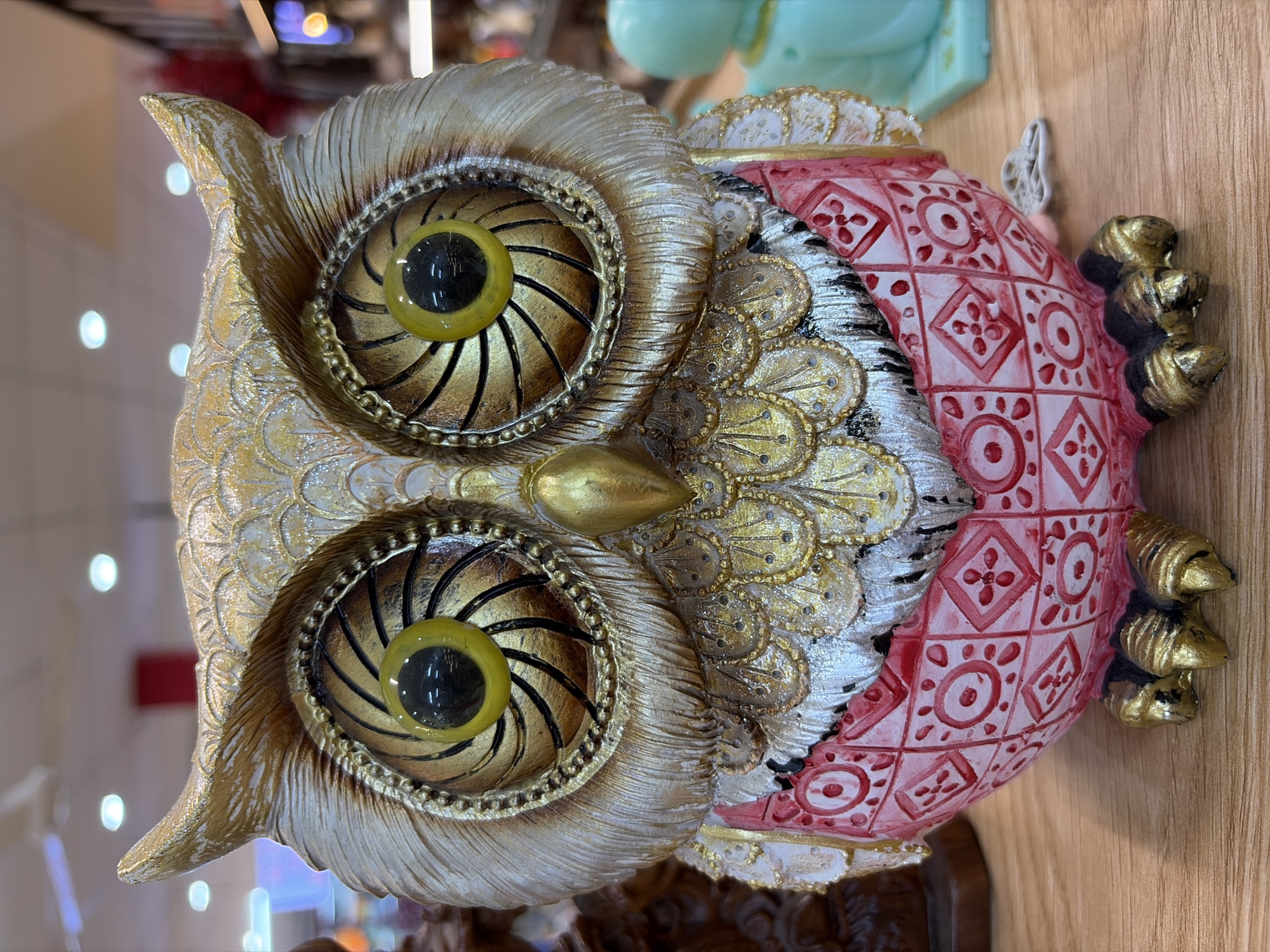 A close-up, eye-level shot shows a decorative owl figurine made of resin. The figurine is gold and white on its head and chest, with large, round, yellow eyes that have black pupils. The owl's body is a reddish-pink with intricate white patterns of circles and diamonds. Its talons are gold. The figurine sits on a wooden surface. In the background, out of focus, are various retail items, including shelving, lights, and other decorative objects. The overall scene suggests a store environment in Da Nang, Vietnam.