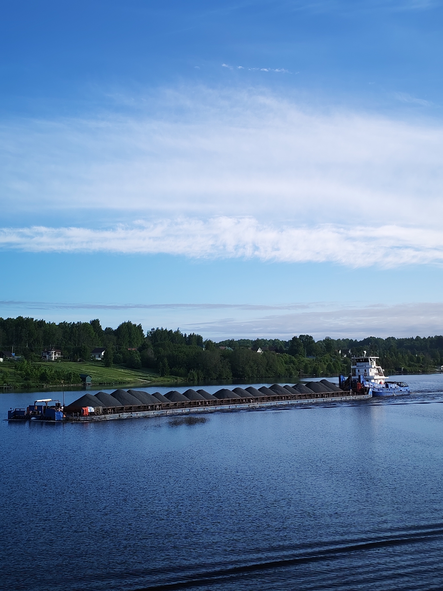 A long barge filled with dark-colored material is being pushed by a white and blue tugboat on a wide, calm river in Раменье, Russia. The barge is nearly parallel to the viewer. The river reflects the blue sky, which has wispy white clouds. The opposite bank is covered in lush green trees and scattered houses. The weather appears calm and sunny. Ripples in the water indicate the barge's movement.
