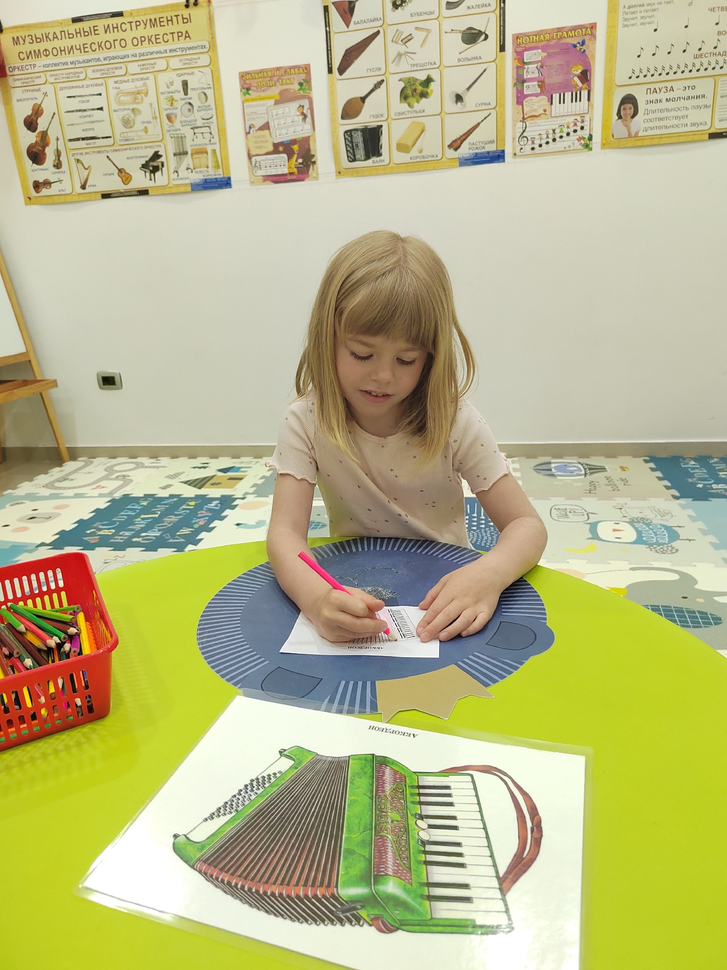 A young girl with blonde hair is seated at a bright green table, intently coloring a musical instrument diagram with a pink pencil. She wears a light-colored, short-sleeved top. To her left, a red basket overflows with numerous colored pencils. On the table in the foreground, a laminated sheet displays an illustration of a green accordion, labeled "АККОРДЕОН" (Accordion).
The scene takes place indoors, likely a classroom or learning center in Podgorica, Montenegro. The walls behind her are covered with educational posters in Russian. One large poster is titled "МУЗЫКАЛЬНЫЕ ИНСТРУМЕНТЫ СИМФОНИЧЕСКОГО ОРКЕСТРА" (Musical Instruments of the Symphony Orchestra), listing instrument categories such as string, woodwind, brass, and percussion, with accompanying illustrations of instruments like the cello, balalaika, and bagpipes. Other posters include "СИЛЬНАЯ И СЛАБАЯ ДОЛИ ТАКТ" (Strong and Weak Beats of a Measure) and "НОТНАЯ ГРАМОТА" (Music Literacy), which features a piano keyboard and musical notes. A poster further to the right explains musical pauses: "ПАУЗА - это знак молчания. Длительность паузы соответствует Длительности звука." (PAUSE - this is a sign of silence. The duration of a pause corresponds to the duration of a sound.)
The floor is covered with colorful, interlocking foam play mats featuring letters ("ABCDEF GHIJKL MNOPQR STUV WXYZ"), numbers, and cartoon animals, along with English words like "Happy." The overall impression is one of a children's learning environment, with the girl actively engaged in a music-related craft.