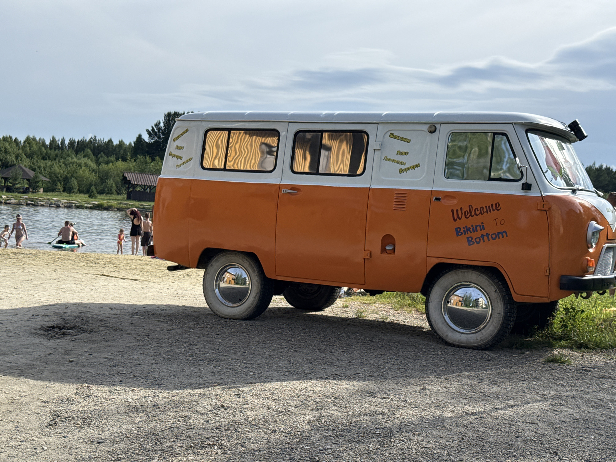 The image captures a beach scene in Syrostan, Russia, featuring an orange and white van parked on a gravel area next to a body of water. The van has text on its side that reads "Welcome Bikini To Bottom." Several people are visible near the water, some wading in it, and one person is on a paddleboard. The background consists of trees and a partly cloudy sky. The overall environment suggests a recreational setting, likely a beach or lake resort.