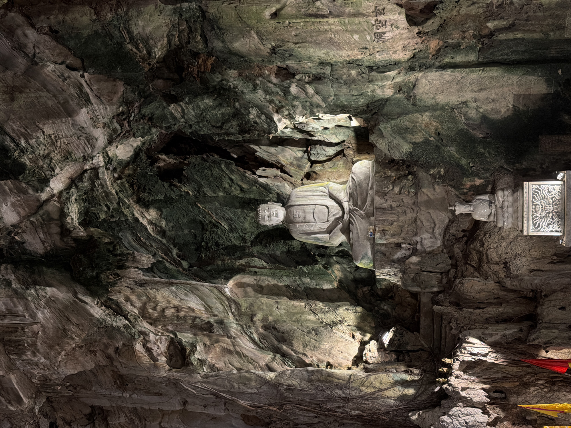 A large, serene Reclining Buddha statue is prominently featured within a natural cave. The Buddha, carved from light-colored stone, lies on its side, with its head resting on a raised platform. The drapery of its robes is intricately detailed, and an auspicious swastika symbol is clearly visible on its chest.
The cave interior is characterized by rugged, uneven rock formations, showcasing varying shades of dark grey, brown, and areas with greenish moss or mineral deposits. Artificial lighting casts dramatic shadows and highlights, emphasizing the textures of the rock and the smooth surface of the statue.
Above the Buddha's head, carved into the cave ceiling, are Chinese characters reading "平安佛" (Píng'ān Fó), which translates to "Peaceful Buddha" or "Buddha of Peace." To the right of the main figure, a smaller, partially obscured statue or carved pillar can be seen. Exposed root systems from overhead vegetation trail across parts of the cave floor and lower walls. In the bottom right corner, small pieces of red and yellow fabric are visible, suggesting offerings or decorations.
The scene conveys a sense of spiritual reverence and the natural beauty of a cave temple, consistent with locations found in Đà Nẵng, Vietnam.