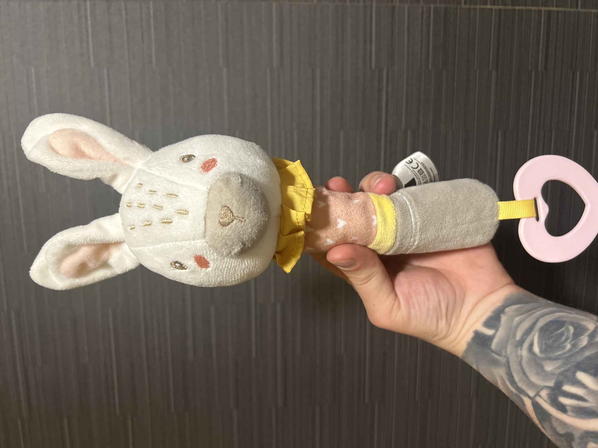 A plush rabbit rattle toy is held in a hand against a dark, vertically-striped wall. The rabbit is mostly white with pink inner ears and a beige nose. It has small embroidered facial features, including red dots for cheeks. Around its neck is a yellow ruffled collar. The rattle handle transitions from pink with white hearts to yellow, then grey. A pink heart-shaped teething ring is attached to the end of the rattle. The hand holding the rattle has a prominent black and white tattoo on the forearm. The location is not identifiable as Varna, Bulgaria, based on visual information.