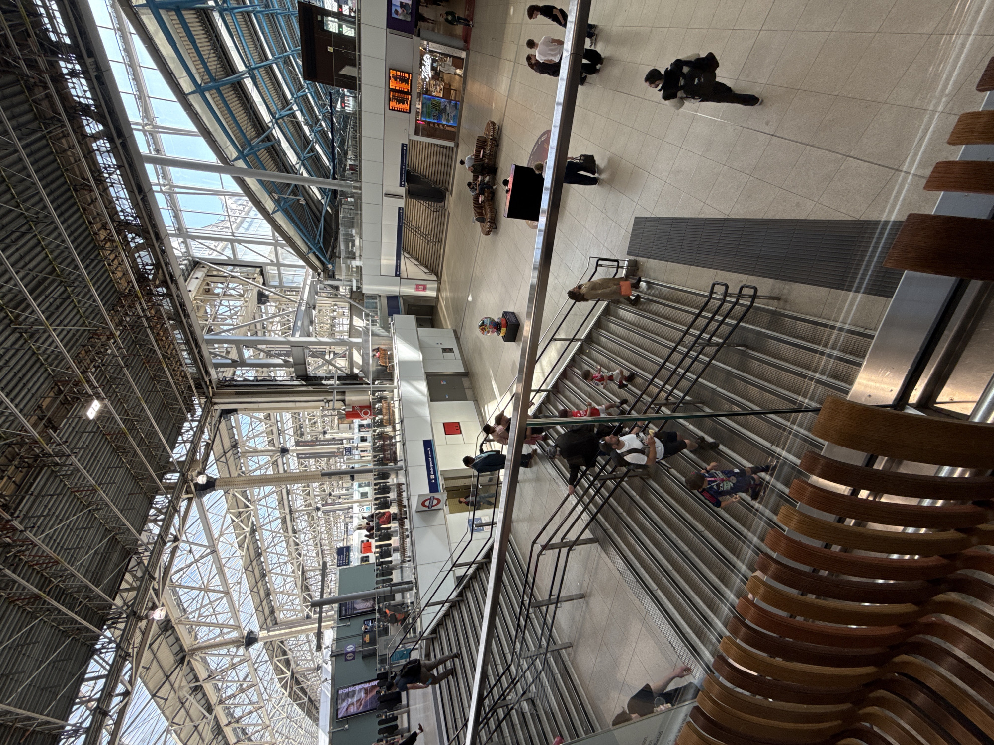 The image depicts an interior view of a train station in London, United Kingdom. The location is likely Victoria Station, as indicated by the signage and architecture. The shot is taken from an elevated position, looking down on the main concourse. The station features a high, vaulted ceiling supported by intricate steel framework with glass panels, allowing natural light to flood the space.
Numerous people are seen walking, standing, and ascending stairs. Some appear to be travelers with luggage. A few individuals are wearing masks.
There are staircases, escalators, and waiting areas with modern seating visible. Signage, digital display boards showing train times, and retail units are present.
The scene appears busy, indicating a peak time at the station. The environment is clean and well-maintained.