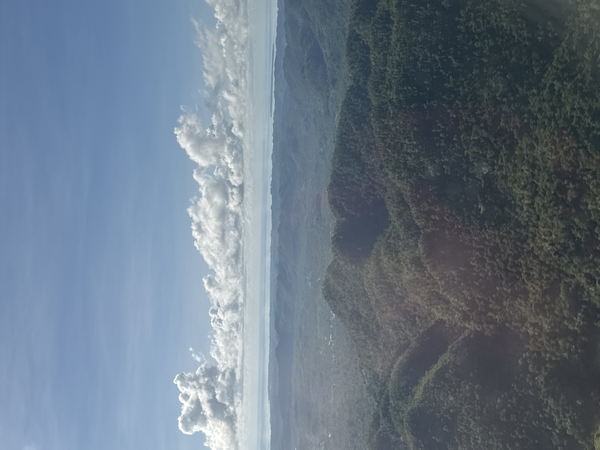 The image shows an aerial view of a landscape with a clear sky and clouds on the left, and a green mountainous terrain on the right. A body of water or horizon line is visible between the sky and the terrain. The landscape appears to be heavily vegetated. While the overall composition suggests a natural environment, without additional context, the specific location within Del Carmen, Philippines, is indistinguishable.