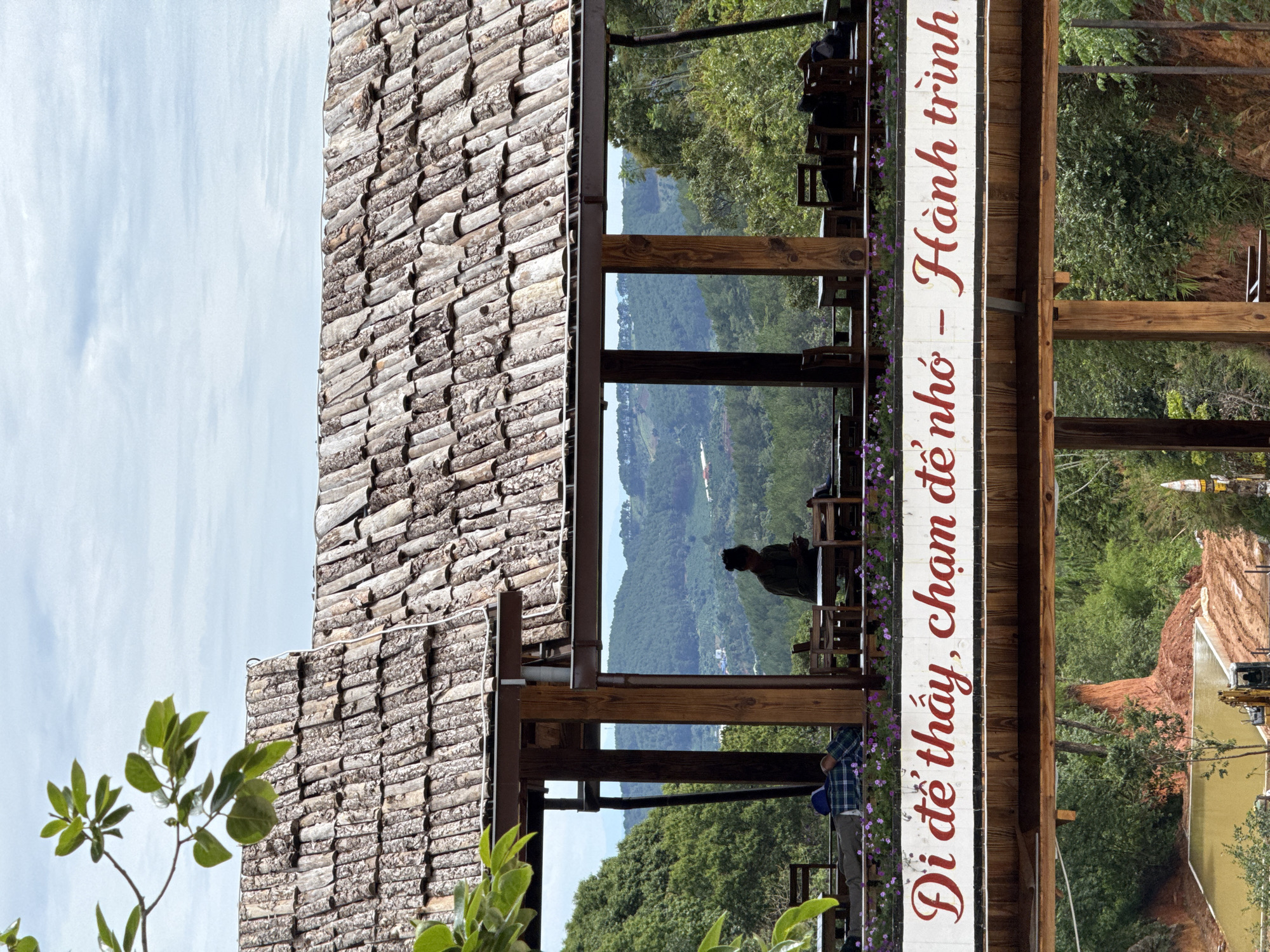 The image depicts a building in Thôn 5, Vietnam, showcasing a unique architectural design. The roof is covered with a pattern of what appears to be closely packed split logs or branches. The building's structure includes an open-air section, likely a porch or dining area, supported by wooden beams. Through the openings, a lush, mountainous landscape is visible, with layered green hills receding into the distance. A person can be seen sitting at a table in the open area, seemingly enjoying the view. A sign, adorned with red cursive Vietnamese text, is positioned prominently on the side of the building. The overall scene evokes a sense of tranquility and connection with nature.