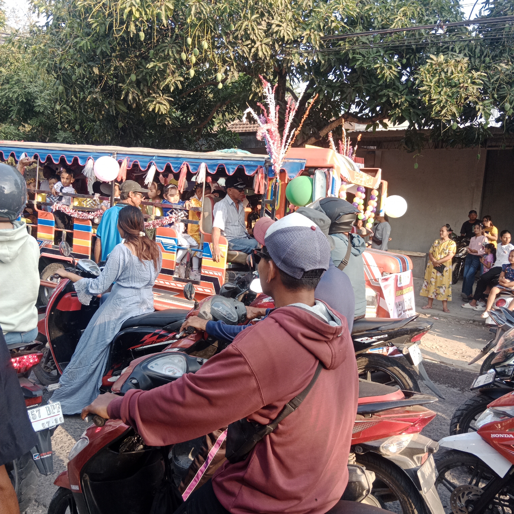 In the city of Tegalrejo, Indonesia, a sunny outdoor scene unfolds with a lively procession of people and vehicles. The focal point is a colorful, open-air transport vehicle, likely a modified truck or bus, filled with passengers. Children and adults are visible inside, some looking out with curiosity. Decorative elements, including colorful streamers and balloons, adorn the vehicle, suggesting a festive occasion.
In the foreground and surrounding the vehicle are numerous motorcycles. Several individuals are seated on these motorcycles, some appearing to be drivers and others passengers. The people are dressed in casual clothing, with many wearing t-shirts, hoodies, and some wearing helmets. The individuals in the foreground are facing away from the camera, with one person in a red hoodie and another in a light blue hoodie.
The environment appears to be a street, with a dirt or gravel surface visible. Tall trees with lush green foliage line the background, casting shadows and indicating a warm climate. Utility lines are also visible crisscrossing above the trees. The overall atmosphere suggests a community gathering or celebration in a typical Indonesian street setting.