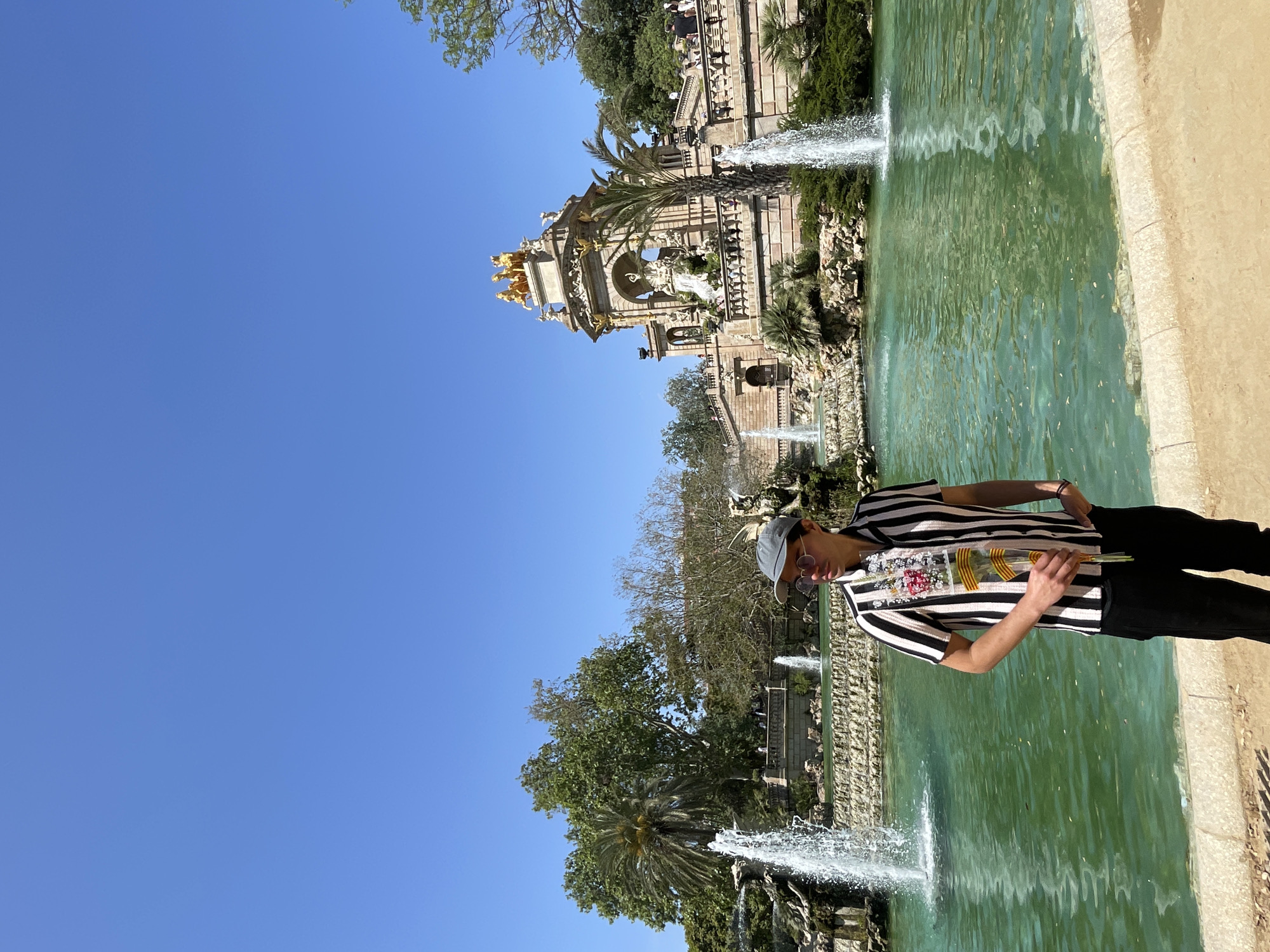 The image shows a person standing beside a fountain in Barcelona, Spain. The person is wearing a black and white striped shirt, black pants, a hat, and sunglasses, and they appear to be holding flowers. The fountain is part of the Parc de la Ciutadella, featuring a large cascading waterfall and ornate architecture with golden statues. The water in the fountain is green in color. The sky is blue.