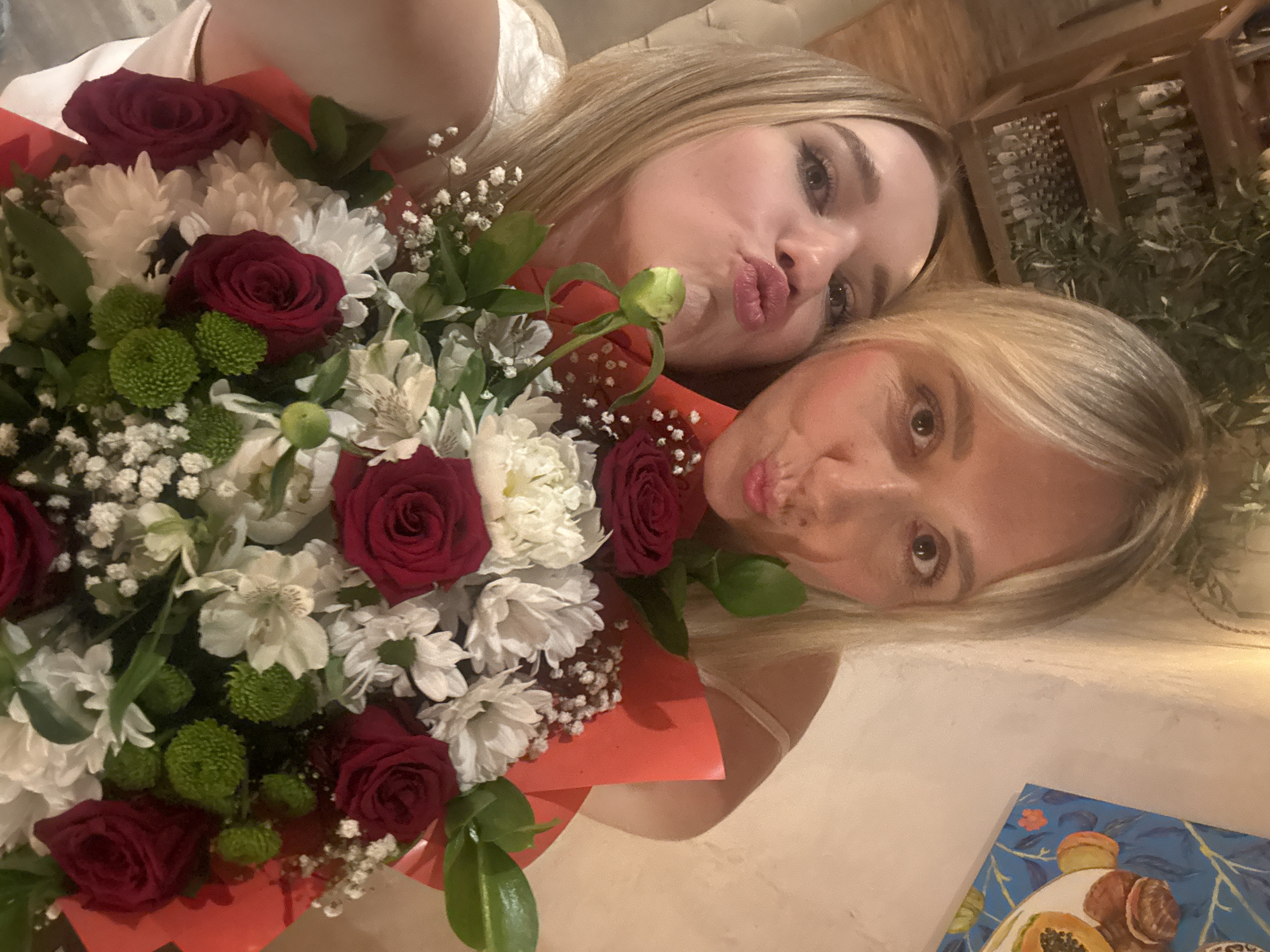 The image shows two women posing for a selfie with a bouquet of flowers. The woman in the foreground is lying down and looking towards the camera with her eyes wide open. The woman in the background is leaning over the bouquet and puckering her lips towards the camera. Both women have blonde hair. The bouquet is large and colorful, with red roses, white chrysanthemums, and green pom-poms. The background appears to be an indoor setting, possibly a restaurant or home, with wooden shelves and decorative items visible. The lighting suggests it is nighttime or indoors with artificial lighting. The overall mood appears celebratory or affectionate. No visible text is present in the image.